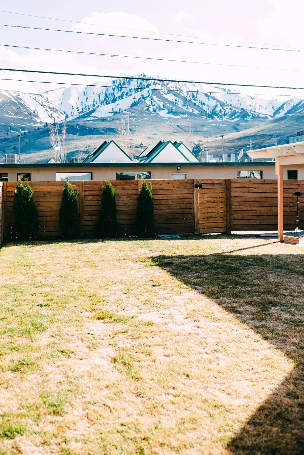 A backyard with a wooden fence, green grass, and trees, with snow-capped mountains in the background under a partly cloudy sky.