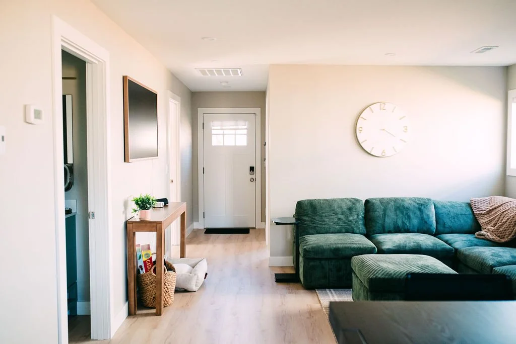 Living room with a green sectional sofa, wall clock, white walls, and a front door with a small window.