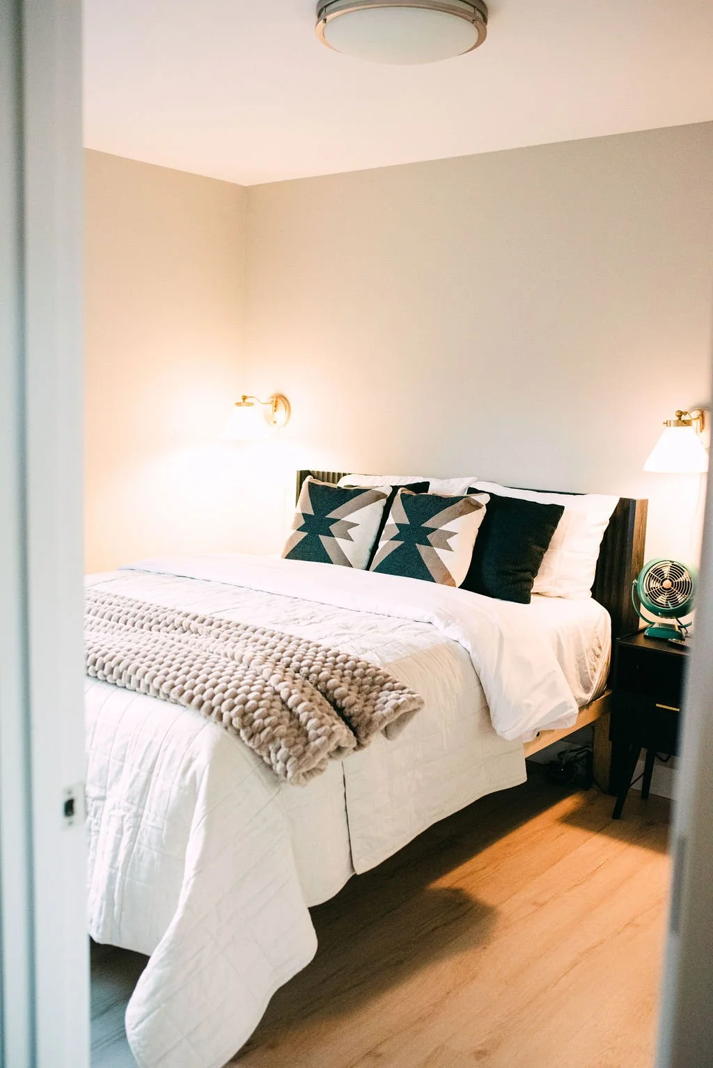 A neatly made bed in a bedroom with beige walls, featuring decorative pillows with geometric patterns, a knitted beige blanket, wall-mounted lamps, a black nightstand with a small fan, and hardwood floors.