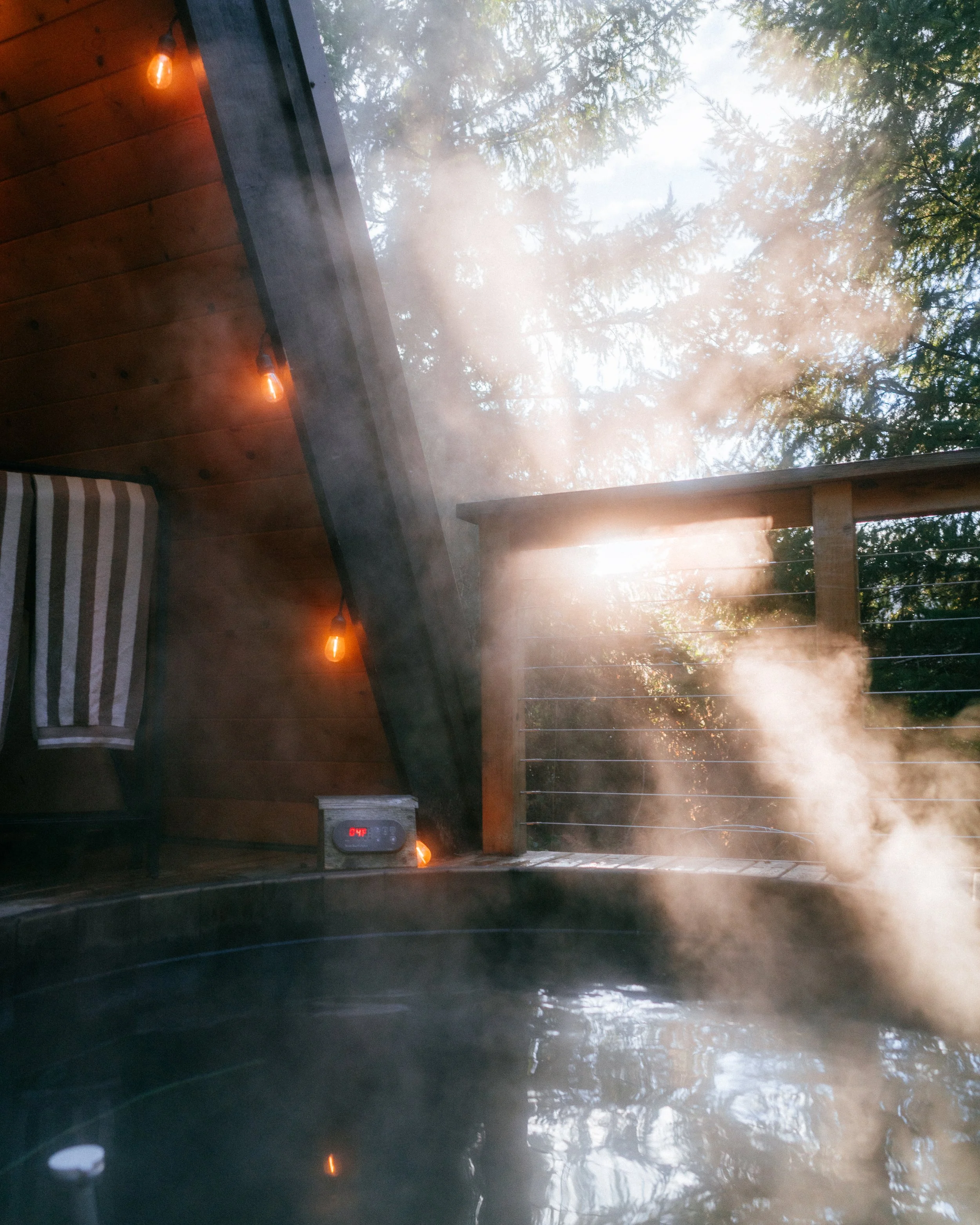 Steam rising from an outdoor hot tub on a wooden deck, with a view of trees and sunlight filtering through the branches.