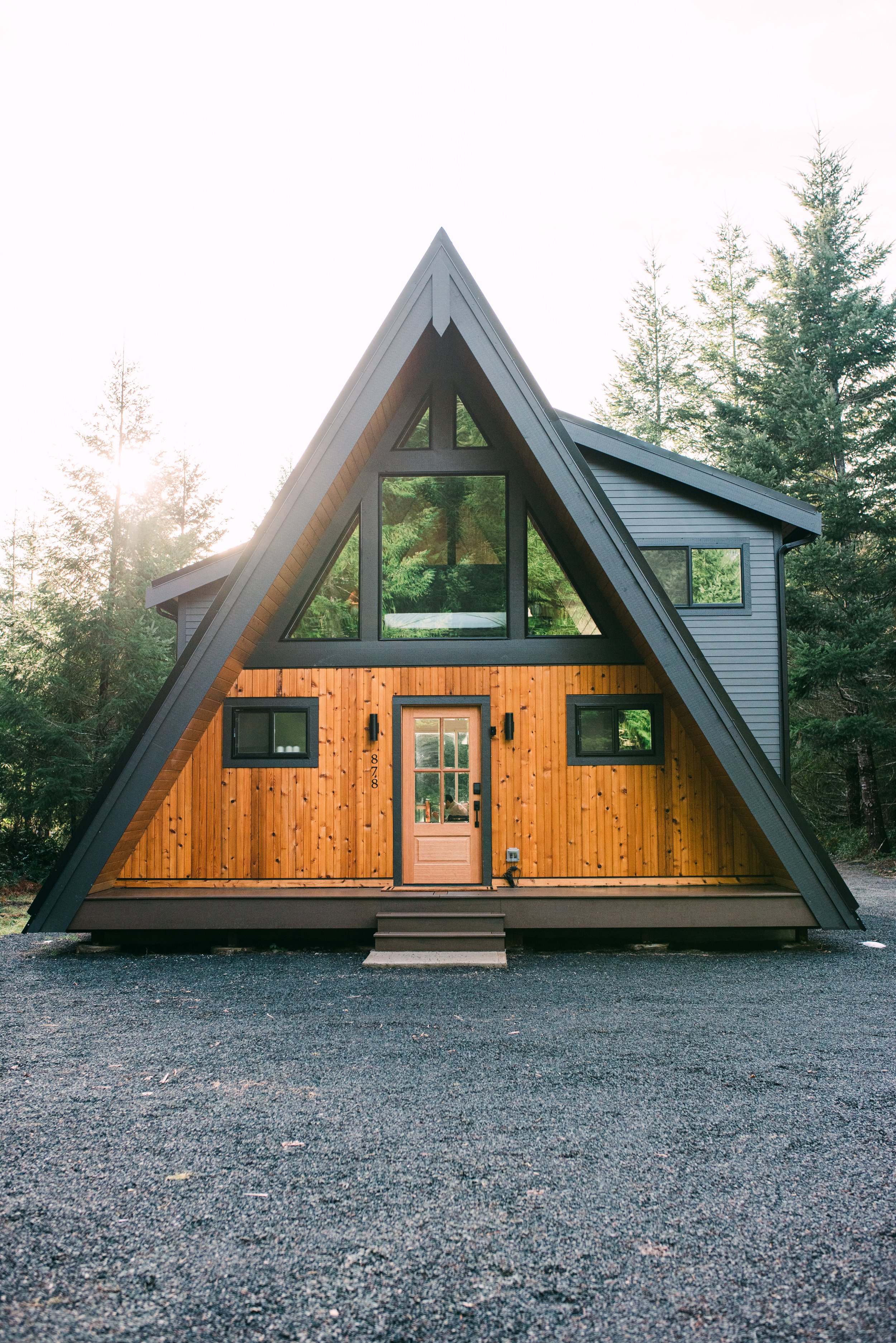 A cozy bedroom with wooden sloped walls and ceiling, featuring a bed with white bedding, black headboard, and two bedside tables with small lamps. A large window and triangular windows provide views of green trees outside, and a woven pendant light hangs from the ceiling.