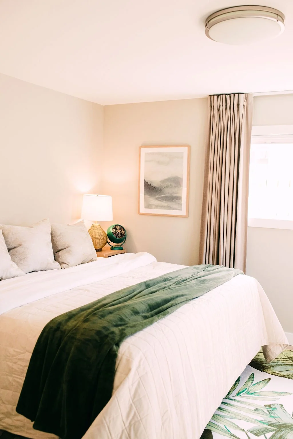 A neatly made bed with beige and gray pillows, a green throw blanket, and a tropical plant-patterned bed skirt. Beside the bed, a wooden nightstand holds a white lamp, a small green fan, and a woven decorative item. Light beige walls feature a framed