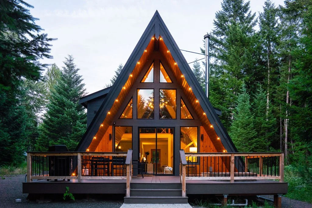 A cozy bedroom with wooden sloped walls and ceiling, featuring a bed with white bedding, black headboard, and two bedside tables with small lamps. A large window and triangular windows provide views of green trees outside, and a woven pendant light hangs from the ceiling.