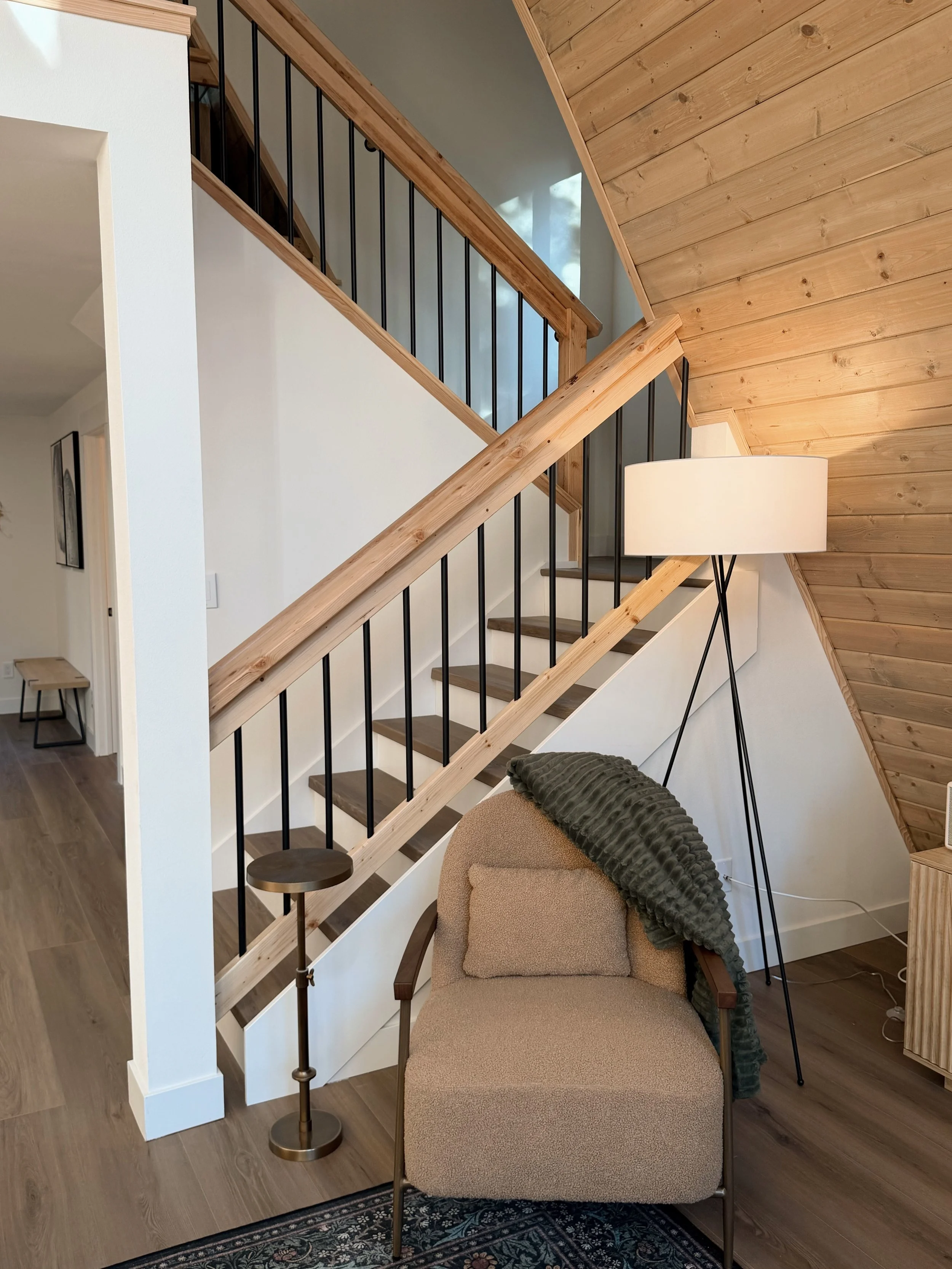 Living room with a staircase featuring wooden steps and black metal railings, a beige armchair with a green throw blanket, a tall white floor lamp, and wooden wall paneling.