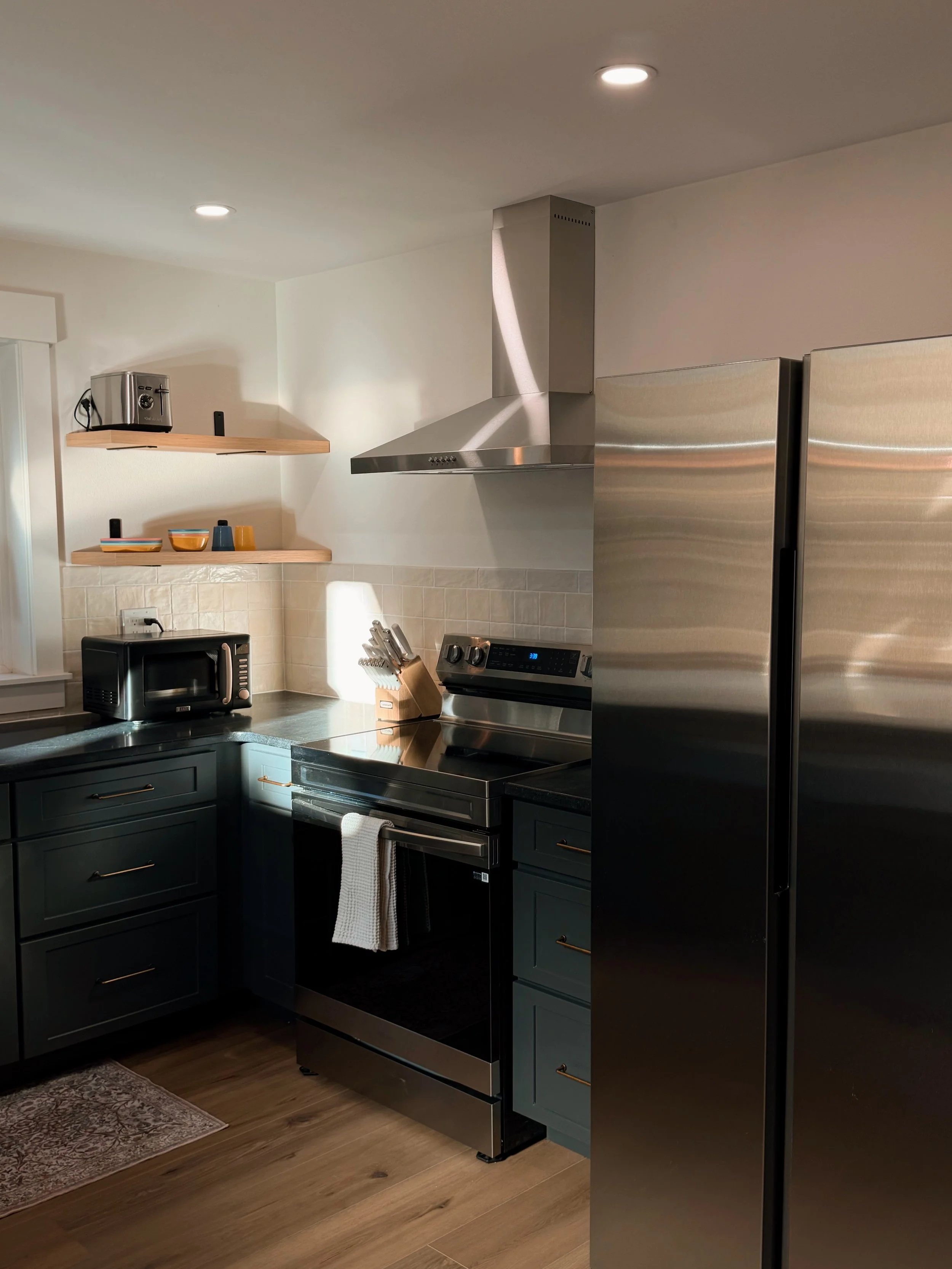 Modern kitchen with black appliances, dark green cabinets, stainless steel refrigerator and range, wooden floating shelves, and hardwood flooring.