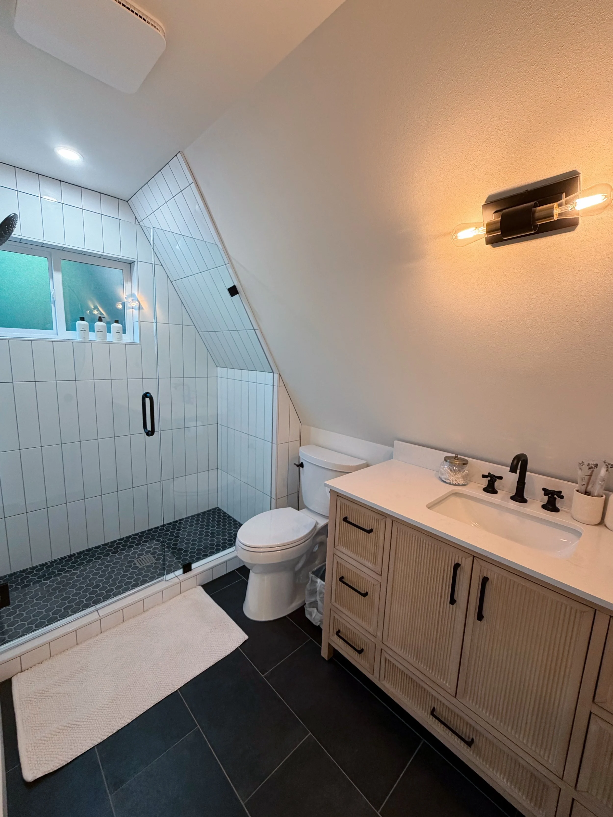 Modern bathroom with a walk-in shower, white tiled walls, a frosted window, a white toilet, a beige vanity with a white countertop, a black faucet, and a black wall-mounted light fixture on a beige wall.
