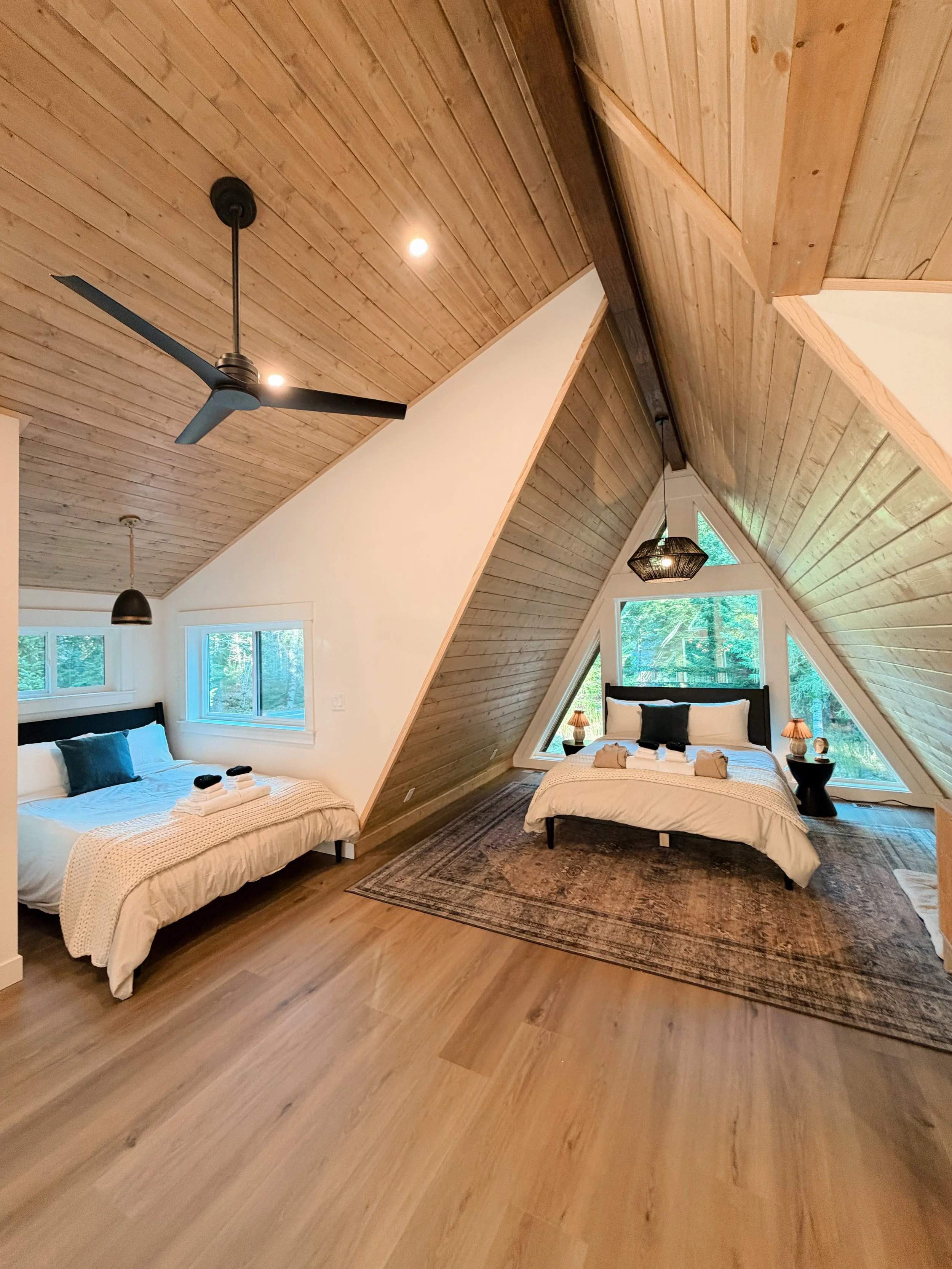 Two bedrooms in an attic with wood-paneled ceilings and walls, each with a bed, nightstands with lamps, and large windows showing greenery outside.