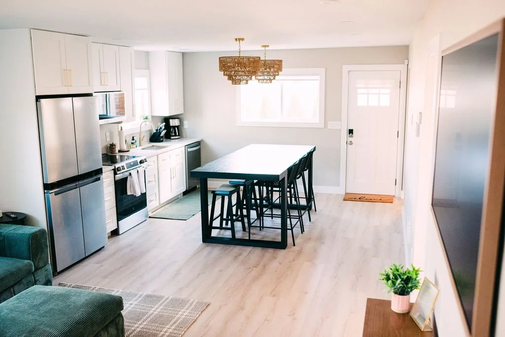 Open-concept kitchen and dining area with white cabinets, stainless steel refrigerator, stove, and microwave, dining table with black chairs, large window, door, wall-mounted TV, and a small side table with potted plant.