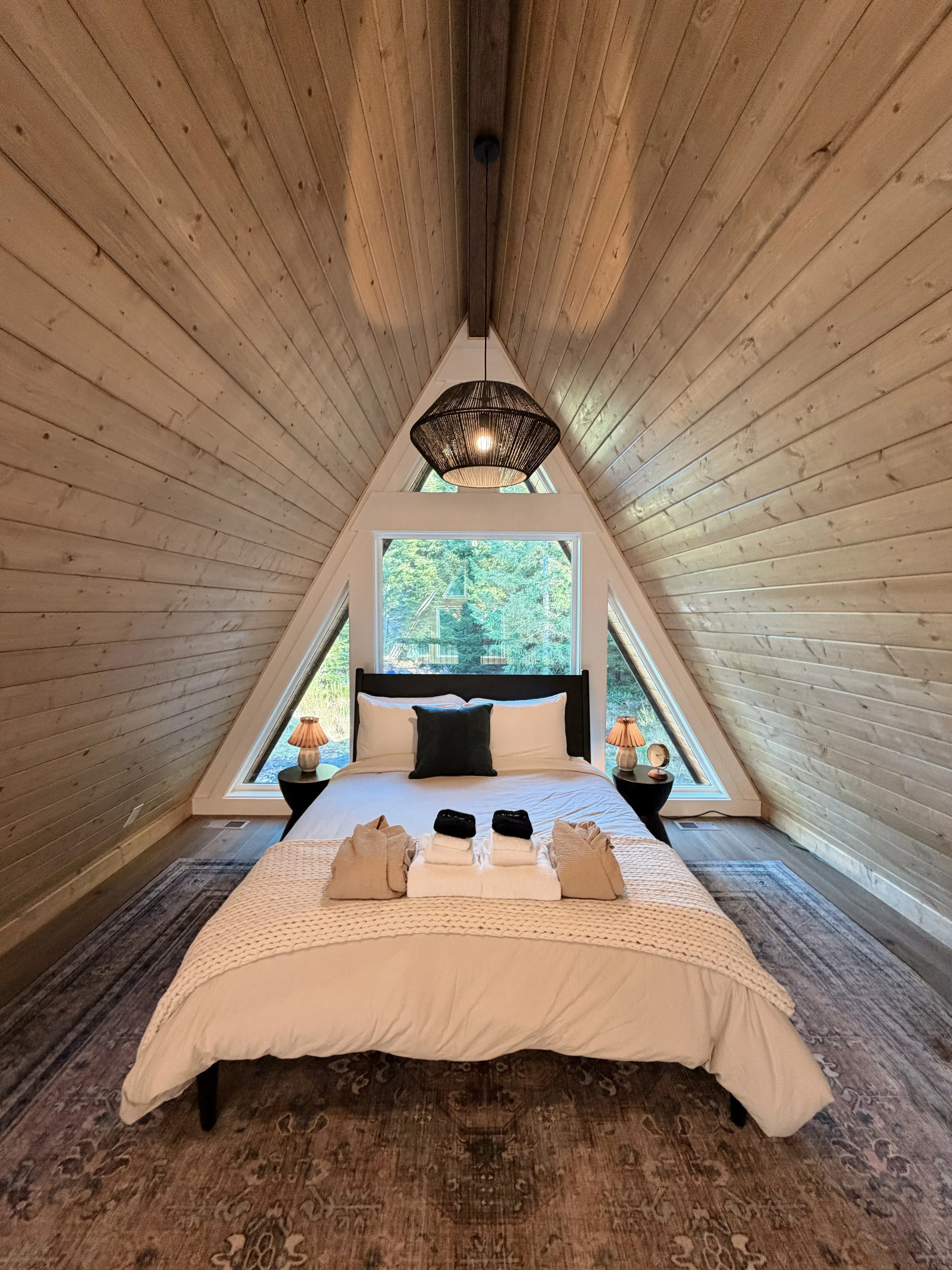 A cozy bedroom with wooden sloped walls and ceiling, featuring a bed with white bedding, black headboard, and two bedside tables with small lamps. A large window and triangular windows provide views of green trees outside, and a woven pendant light hangs from the ceiling.