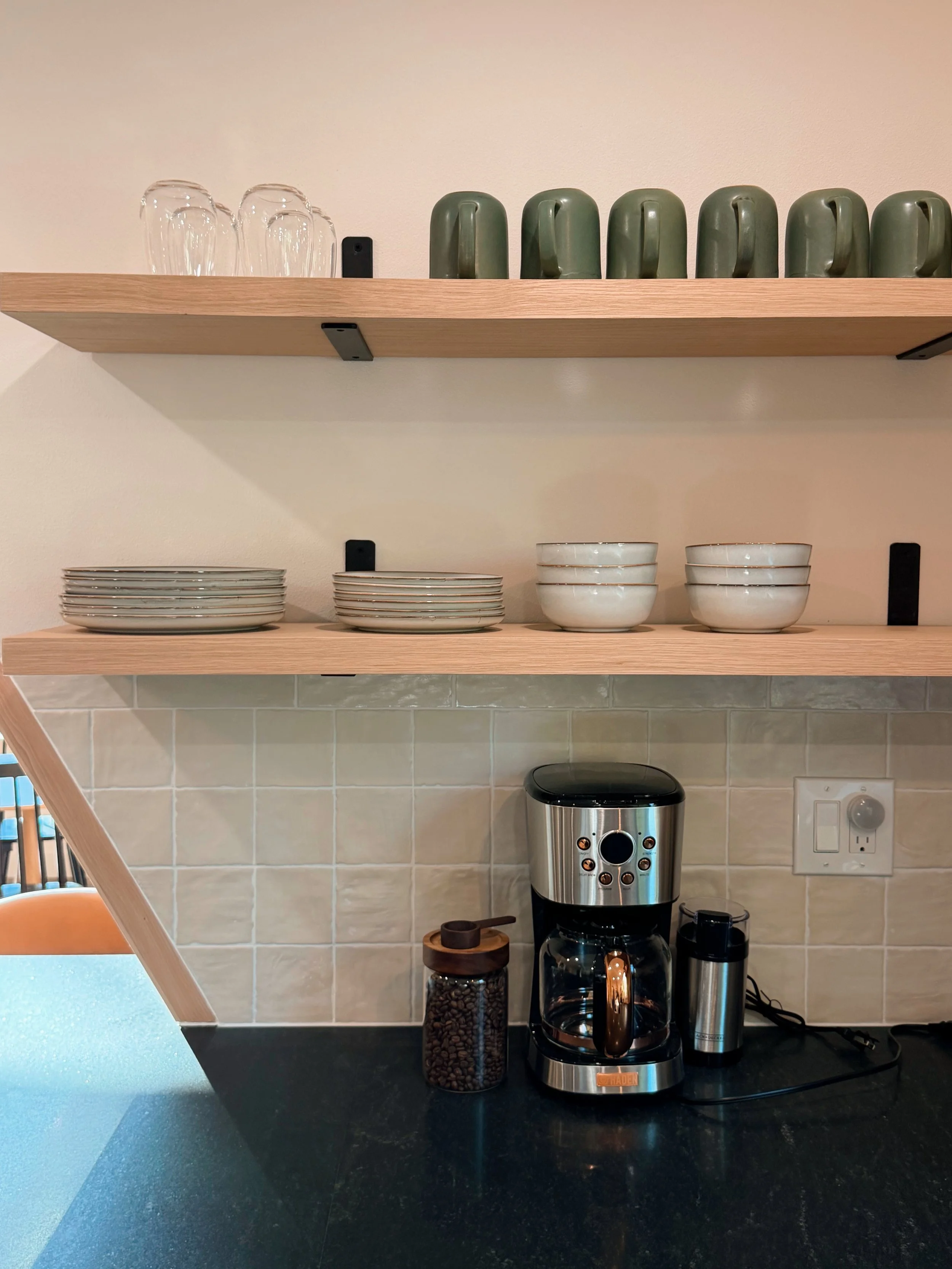 Kitchen with two wooden shelves holding glasses, plates, and bowls; coffee maker, jar of coffee beans, and electric kettle on black countertop.