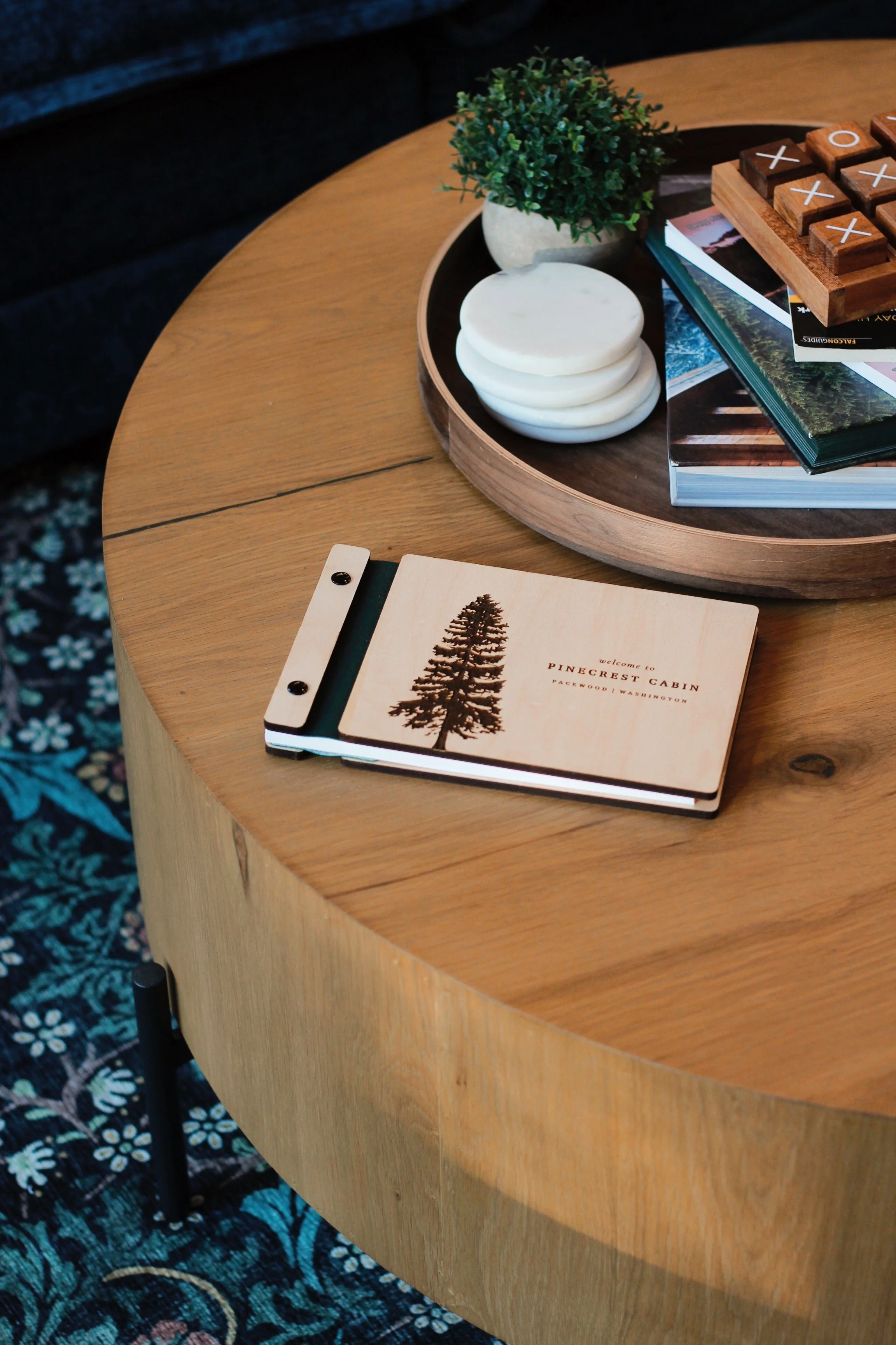 A wooden coffee table with a small black and white notebook displaying a pine tree and the text "Welcome to Pinecrest Cabin, Packwood, Washington," alongside a black pen and a bead with small black dots.