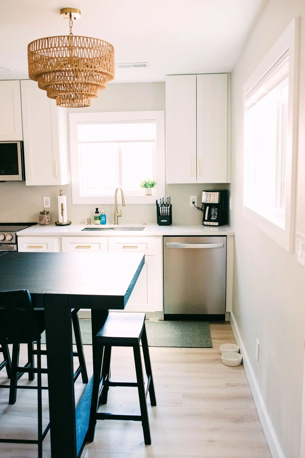 Bright kitchen with white cabinets, a window above the sink, a black countertop island with stools, a woven chandelier, and small appliances including a coffee maker and knife block.