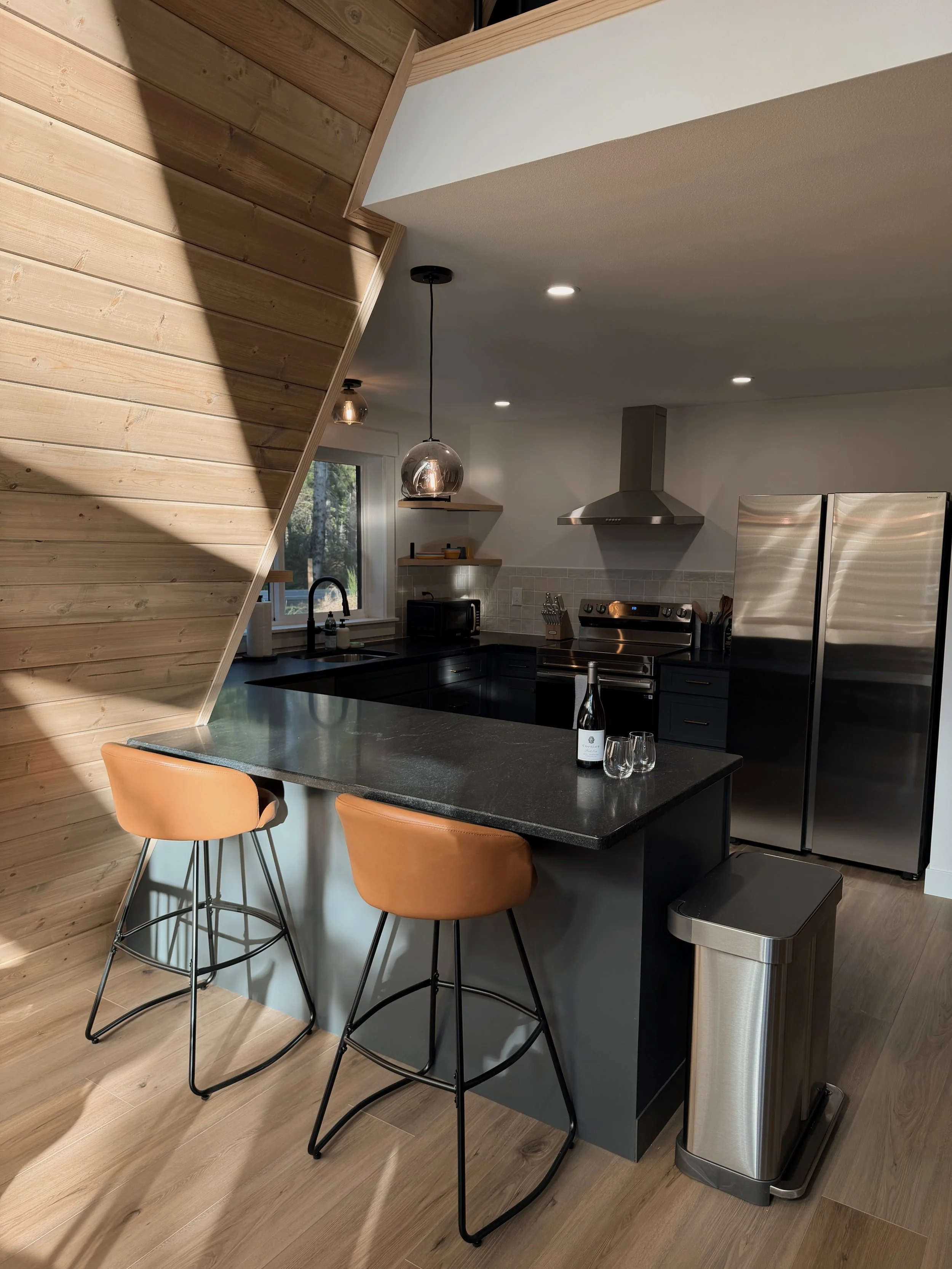 Modern kitchen with black countertops, stainless steel appliances, light wood flooring, and a wooden wall. Two tan barstools are at the kitchen island. There are pendant lights and a bottle with glasses on the counter.