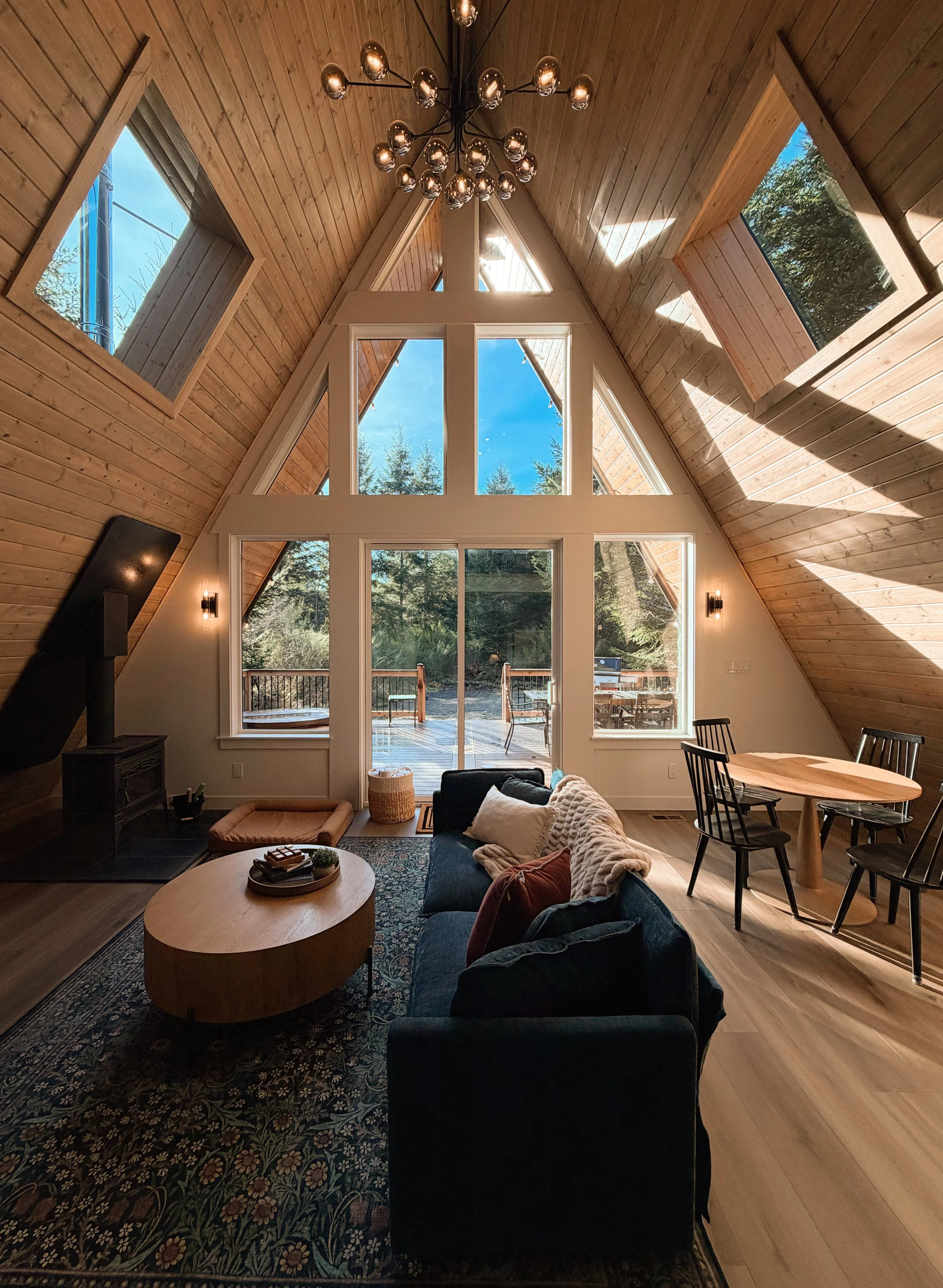 Living room with vaulted wooden ceiling, large triangular windows, and a sliding glass door leading to an outdoor deck. Contains a black sofa with pillows, a round wooden coffee table, a patterned area rug, a dining table with chairs, and a modern ch