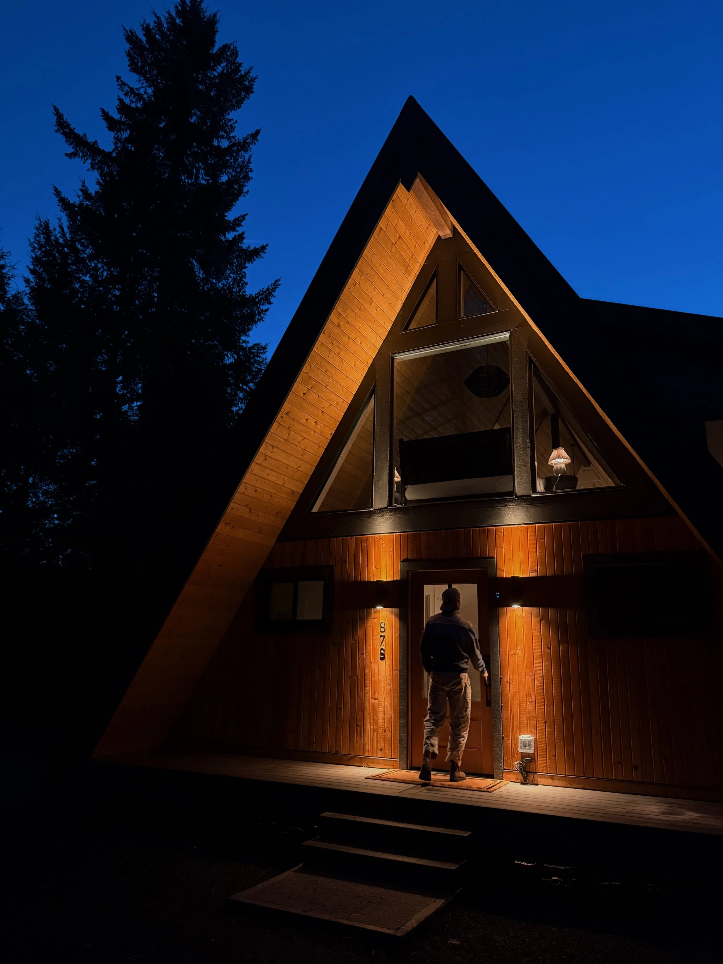 A person standing at the front door of a modern A-frame house at night, with warm lighting illuminating the exterior and a large tree in the background.
