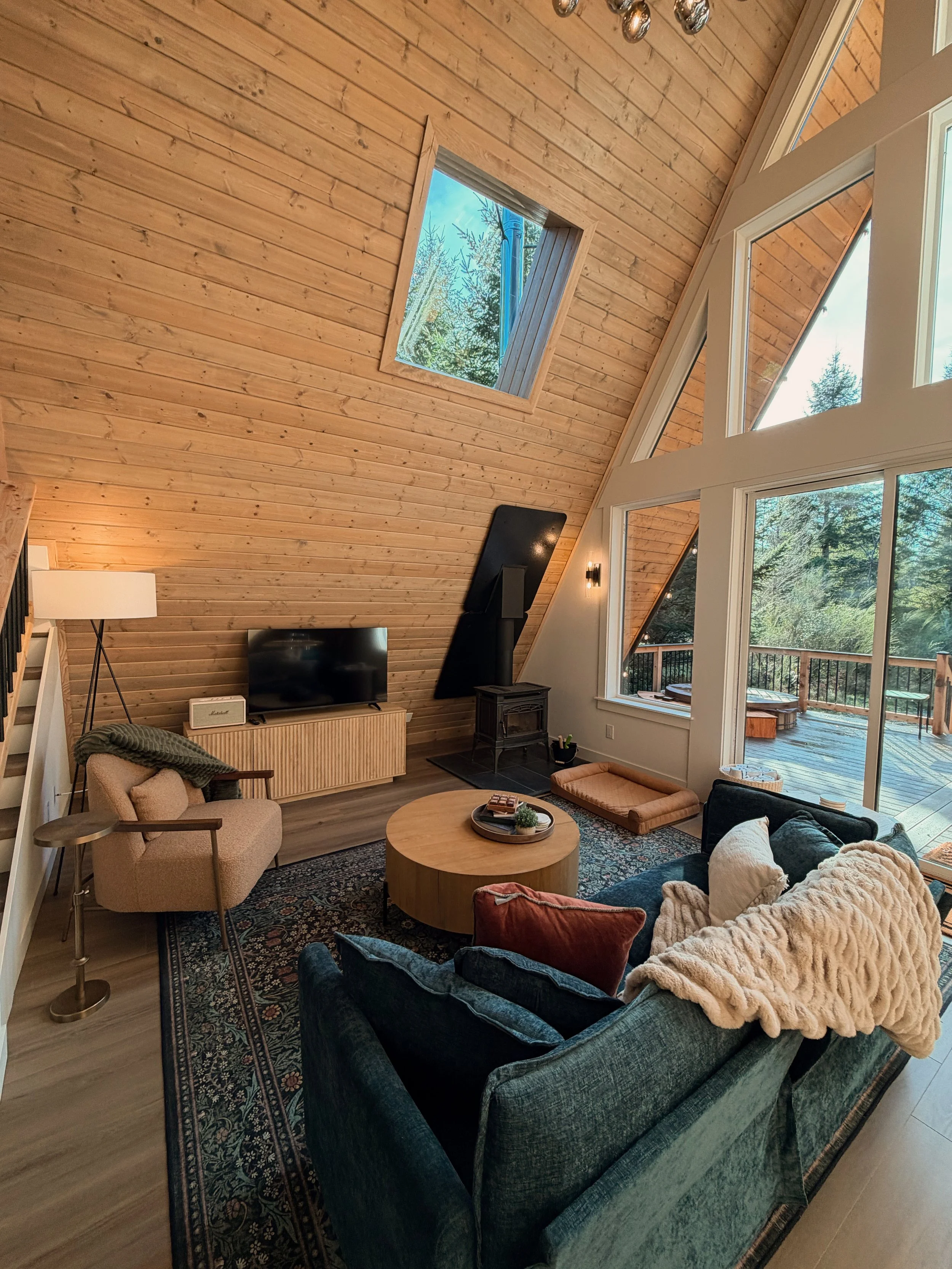 Living room with wood-paneled ceiling, large windows, and a vaulted ceiling with skylight. Includes a dark green sofa, a beige armchair, a round wooden coffee table, a flat-screen TV, and a wood-burning stove.