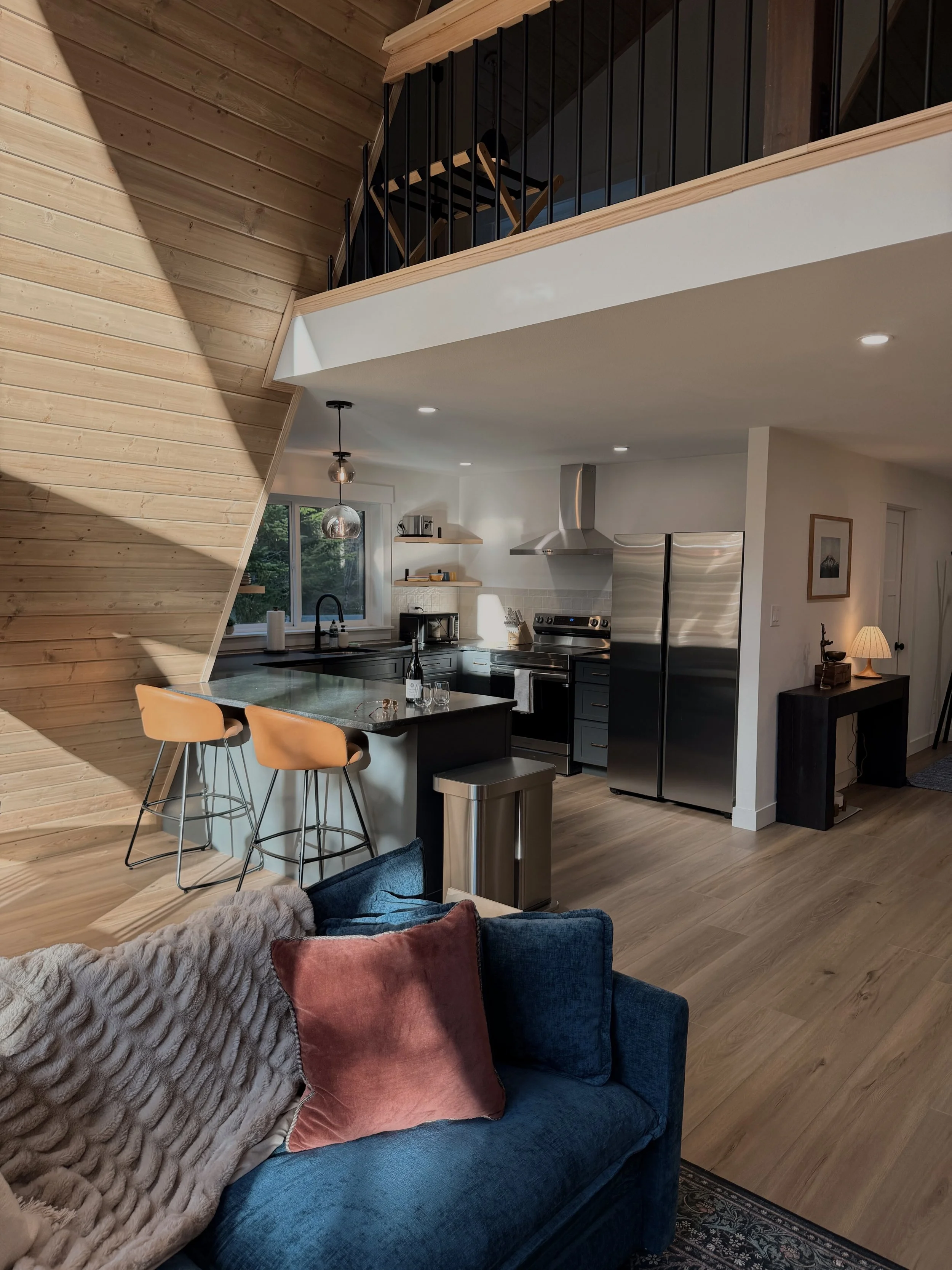 Open-concept living space with a kitchen featuring black cabinets, stainless steel appliances, and wooden open shelves, with a blue sofa and pillows in the foreground.