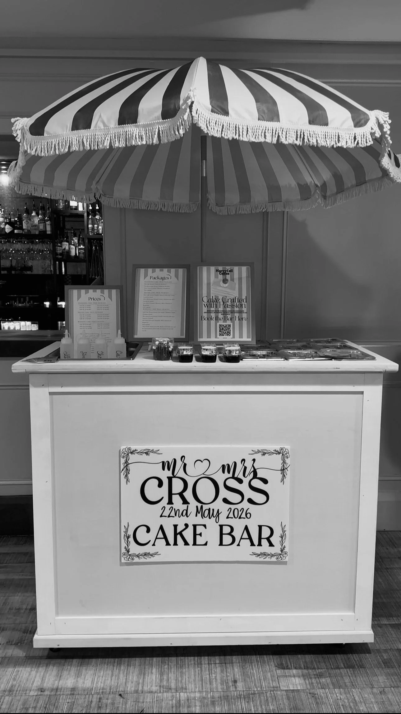 A cake bar booth at a wedding, decorated with a sign that says "Mr & Mrs Cross, 22nd May 2026, Cake Bar." The booth has a large striped umbrella above it, with various cake slices and drinks on the counter.