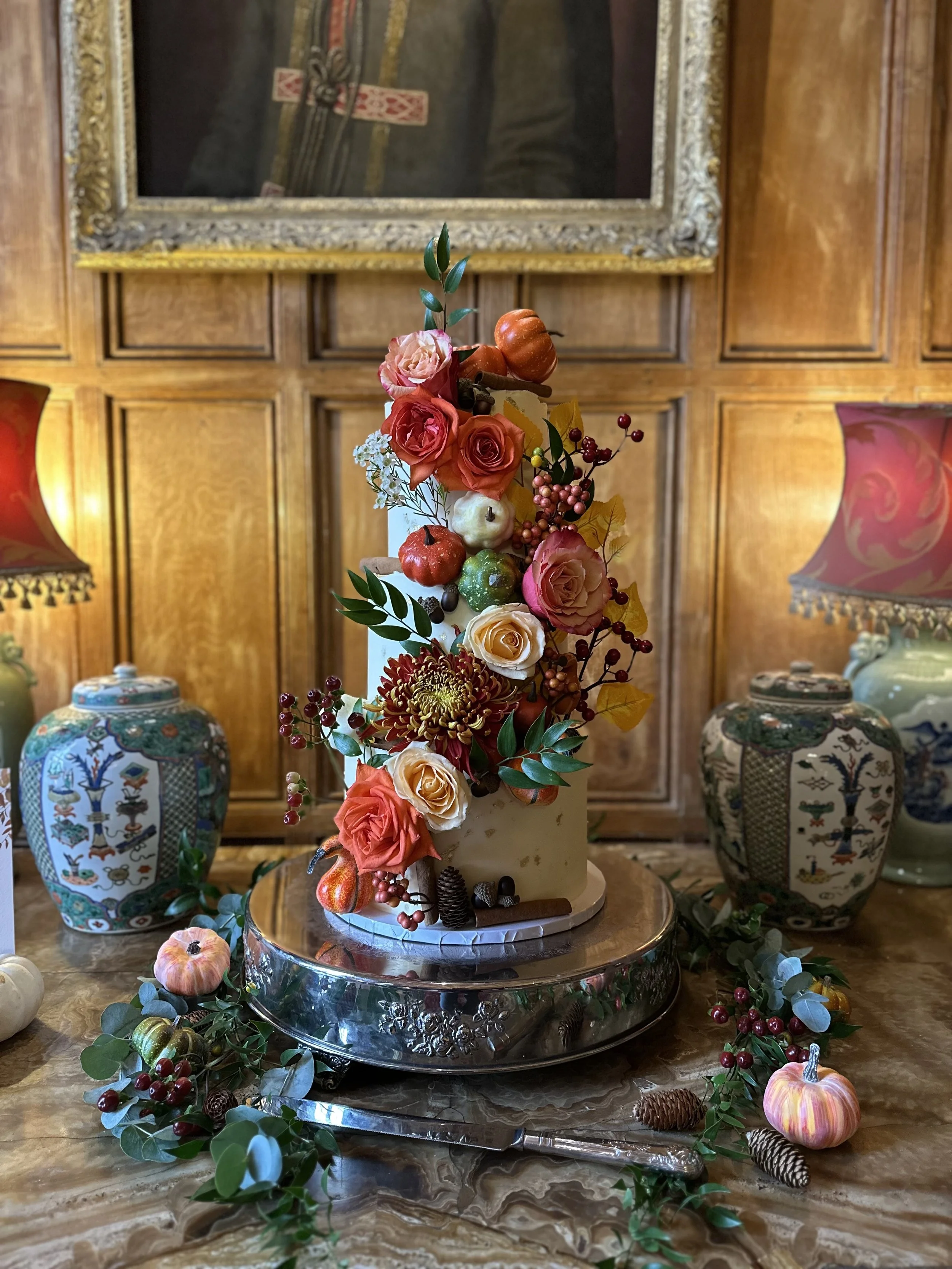 A decorated cake with flowers, apples, and leaves, placed on a silver cake stand on a marble table with fall-themed decorations including small pumpkins, pinecones, and branches.
