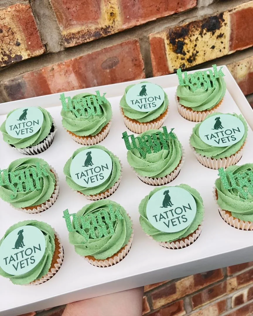 A white tray holding twelve cupcakes decorated with green frosting. Each cupcake has a round topper with a black dog and the text "TATTON VETS". Some cupcakes are topped with glittery green "thank you" signs.