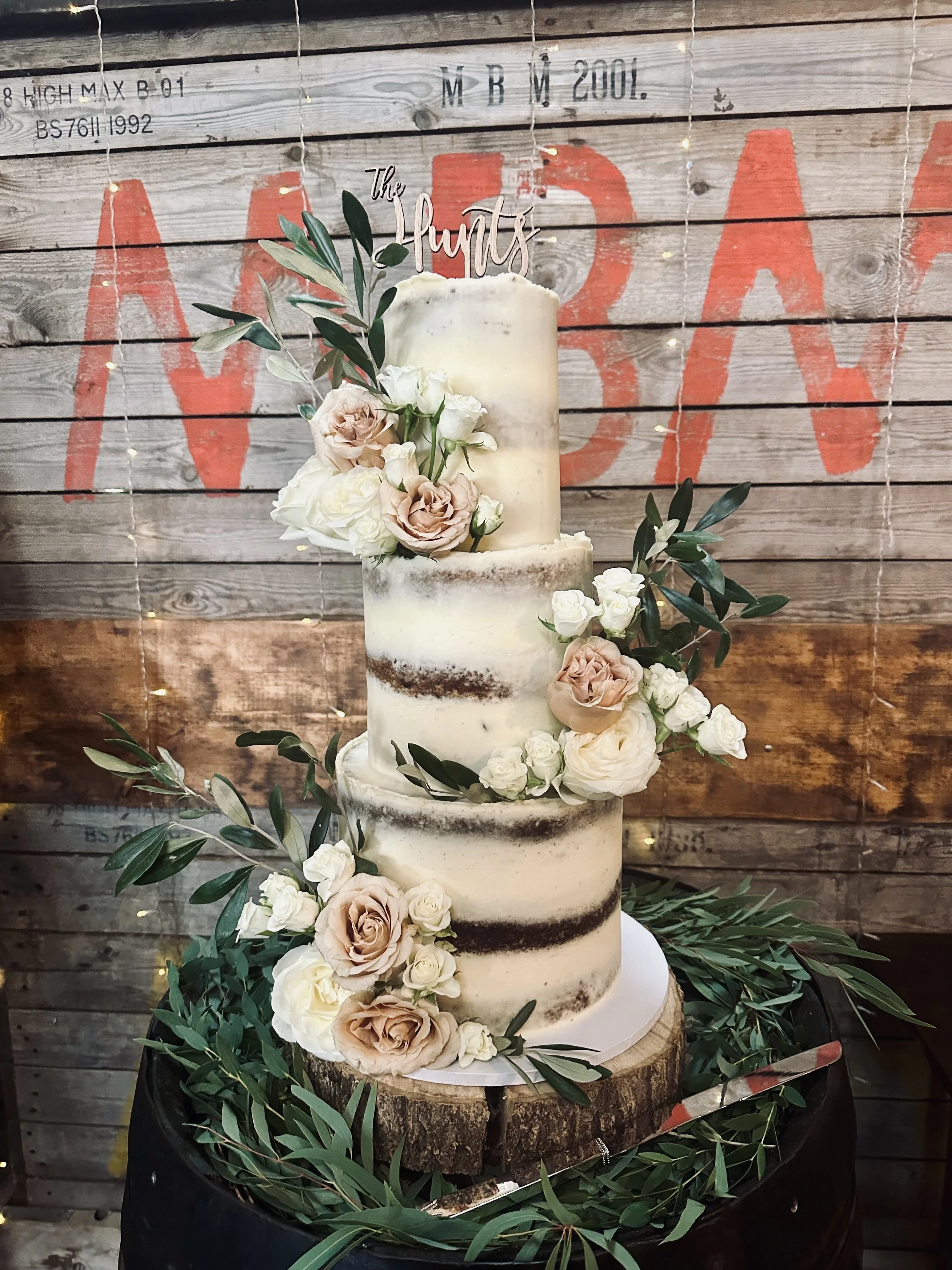 A tall, rustic, semi-naked wedding cake decorated with blush and white roses, green leaves, and a 'The Hunts' cake topper, placed on a wooden slice surrounded by greenery.