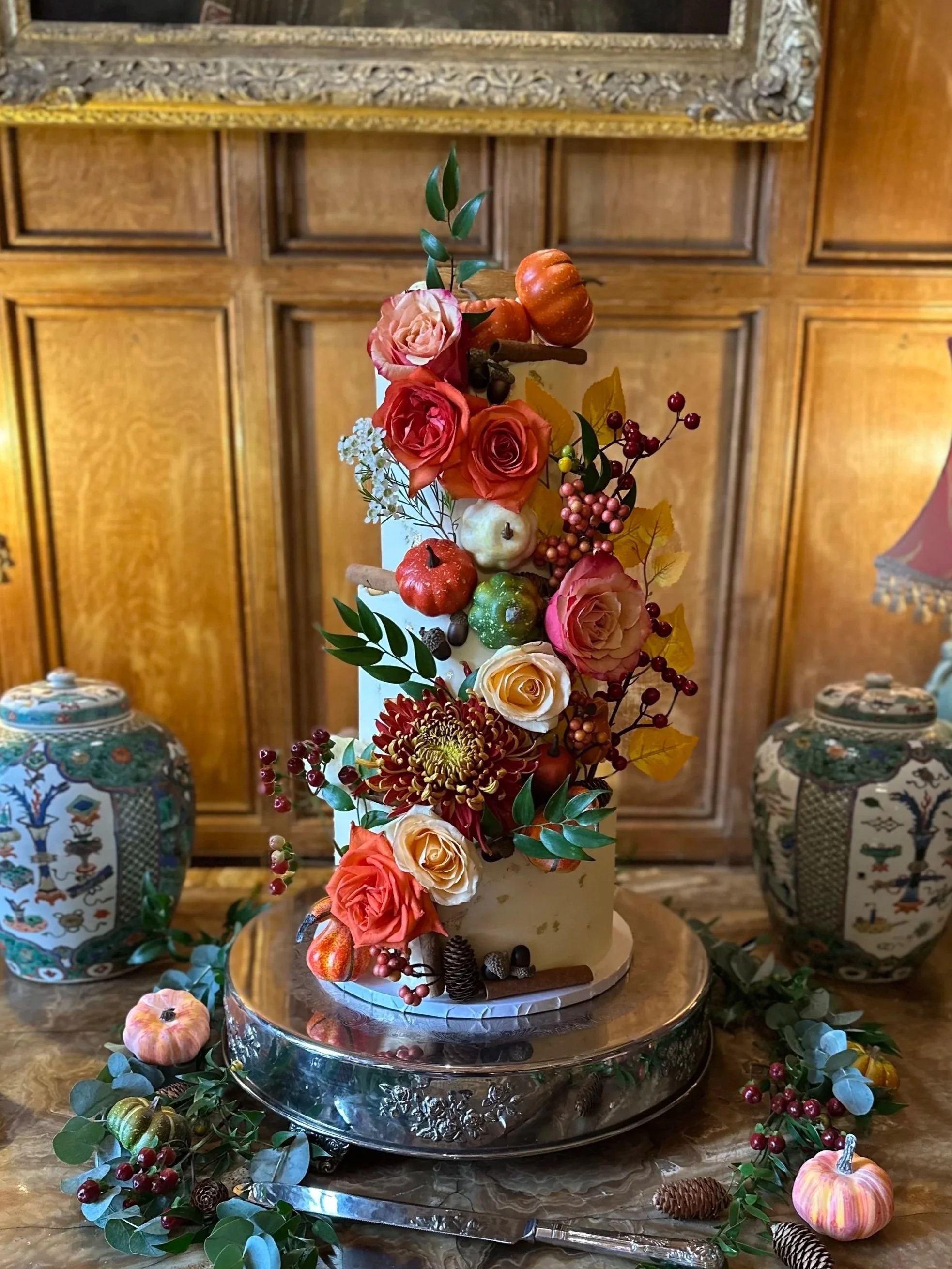 A tall, white cake decorated with various autumn-themed flowers, small pumpkins, apples, berries, and leaves. The cake is displayed on a silver platter with fall decorations around it. In the background, there are wooden paneled walls and decorative porcelain vases.