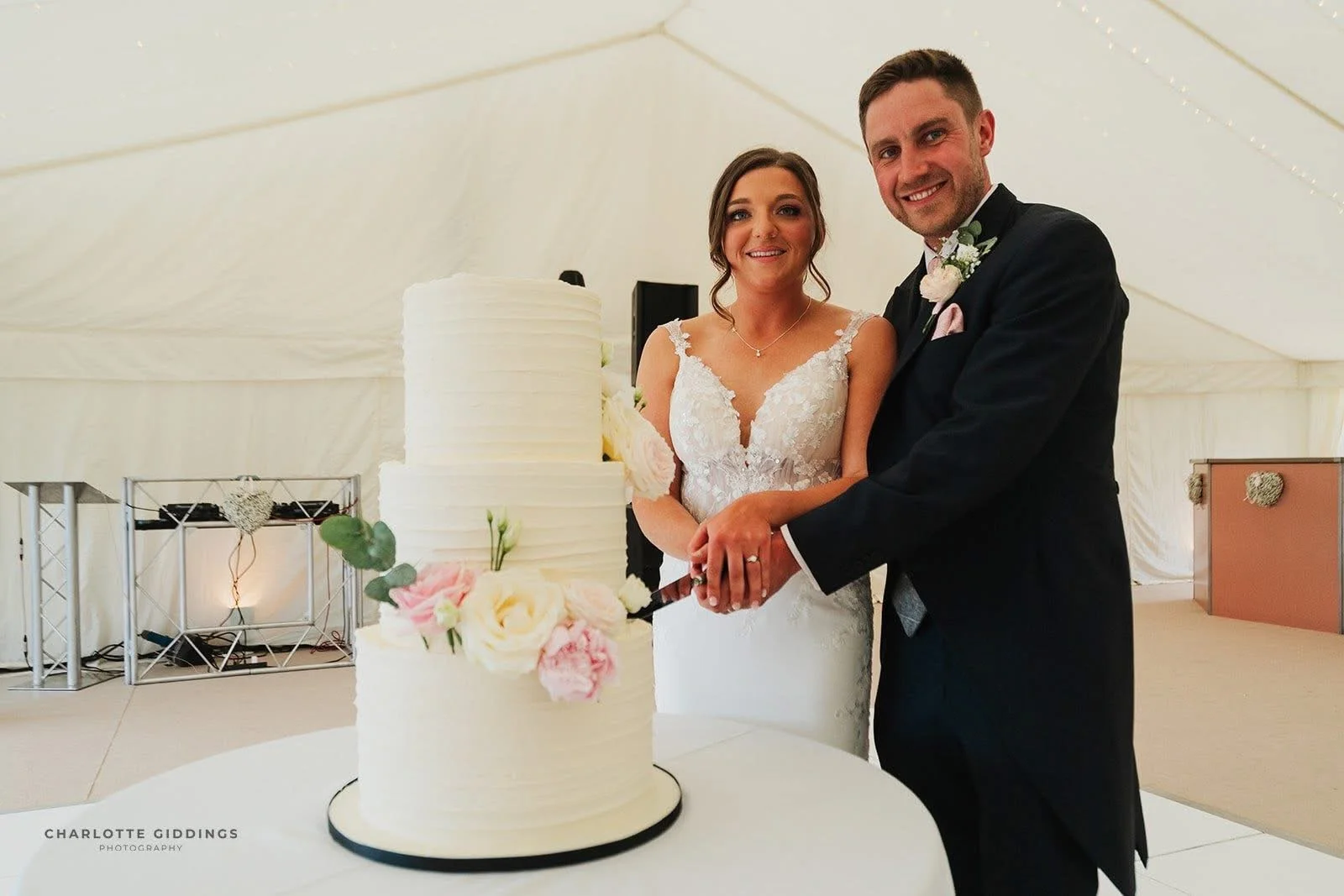 Bride and groom with their three-tier white wedding cake decorated with soft pink and white flowers in Cheshire