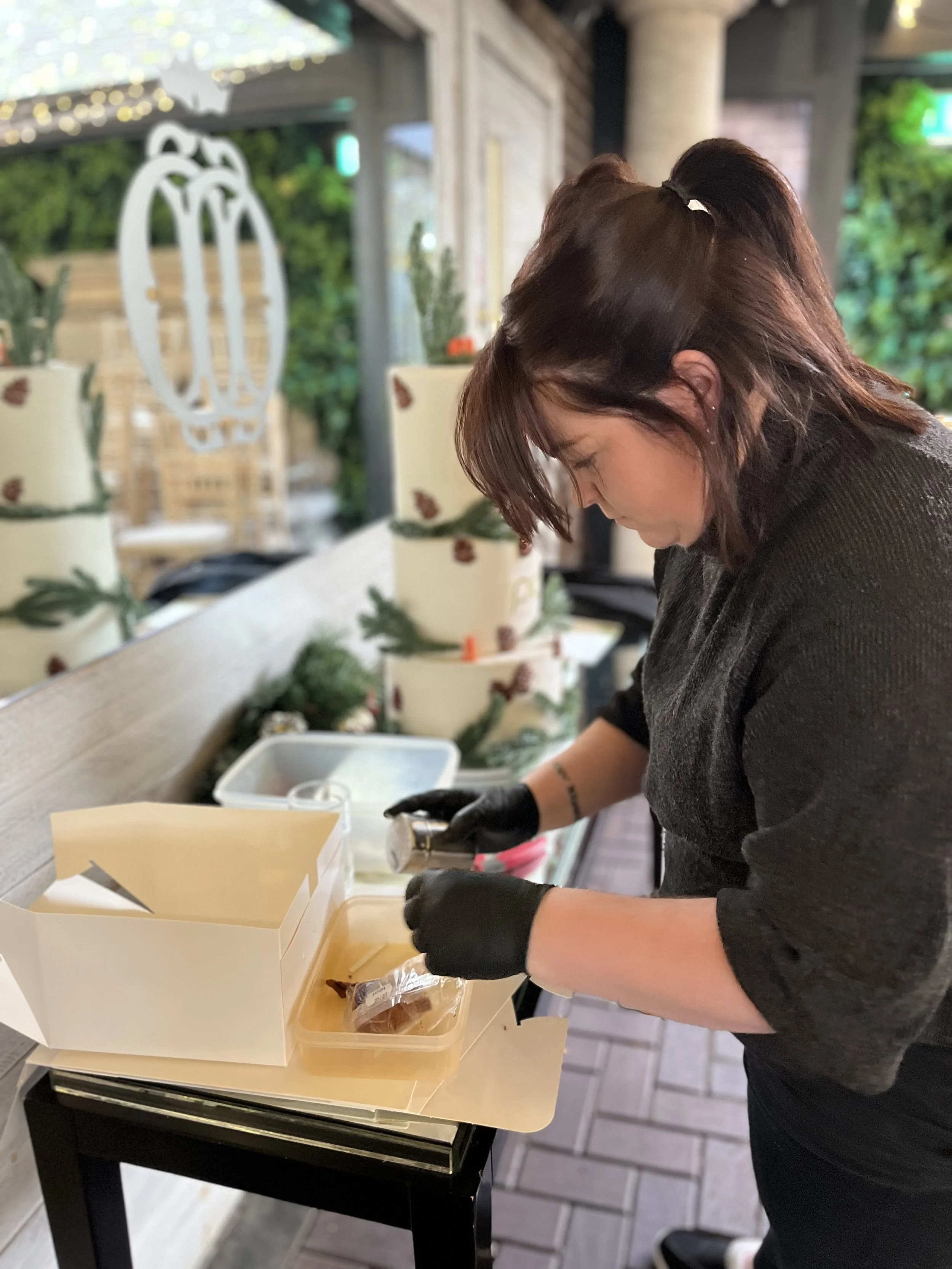 A woman wearing black gloves is packaging baked goods in a white box on a black table inside an indoor space with holiday decorations, including candles and greenery.