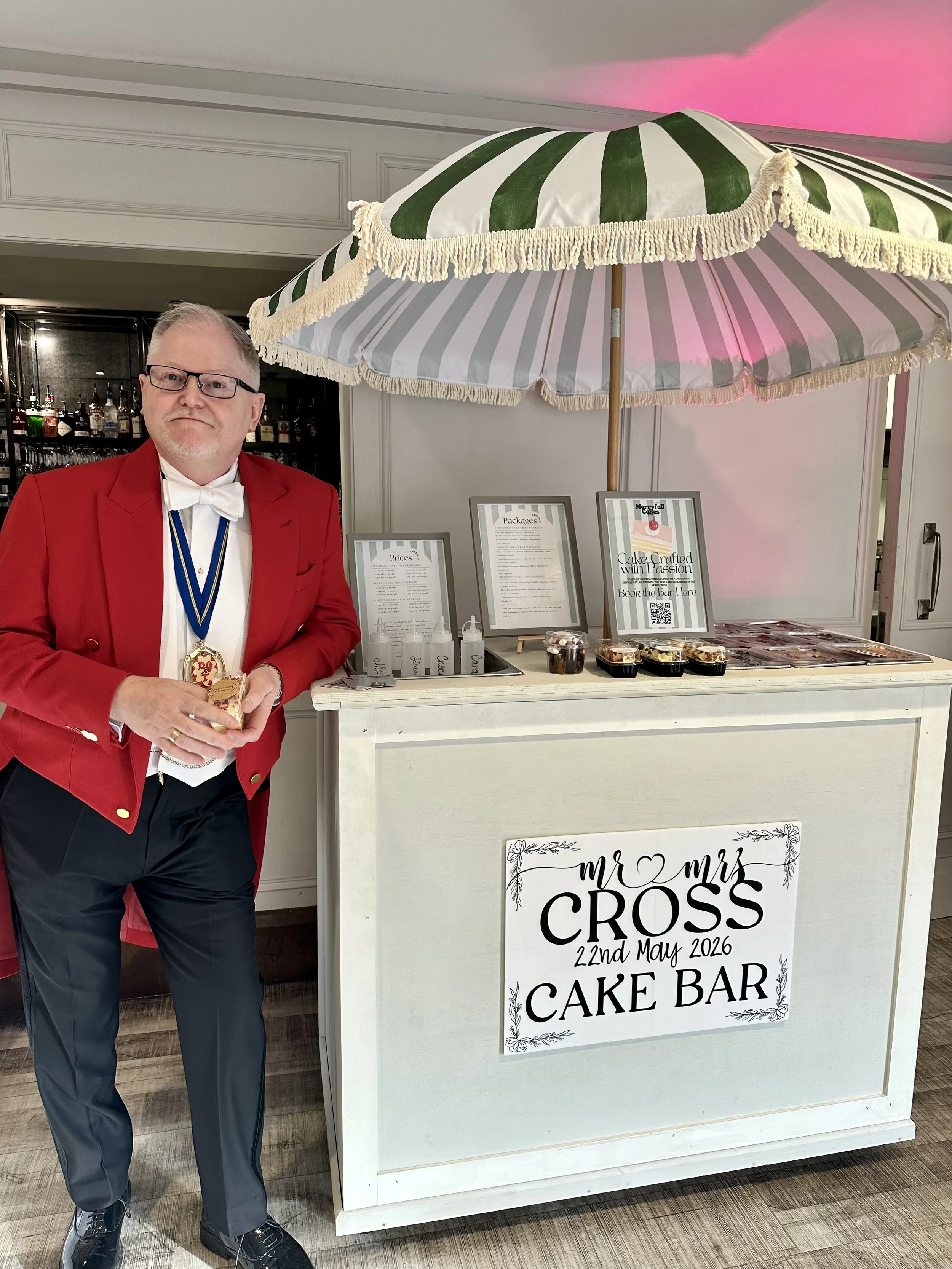 A man in a red jacket, white shirt, and bow tie standing beside a cake bar at a wedding, with a large green and white striped umbrella above the cake bar. The cake bar has a sign that reads 'Mr & Mrs Cross, 22nd May 2026, Cake Bar' and various desserts and menus on display.