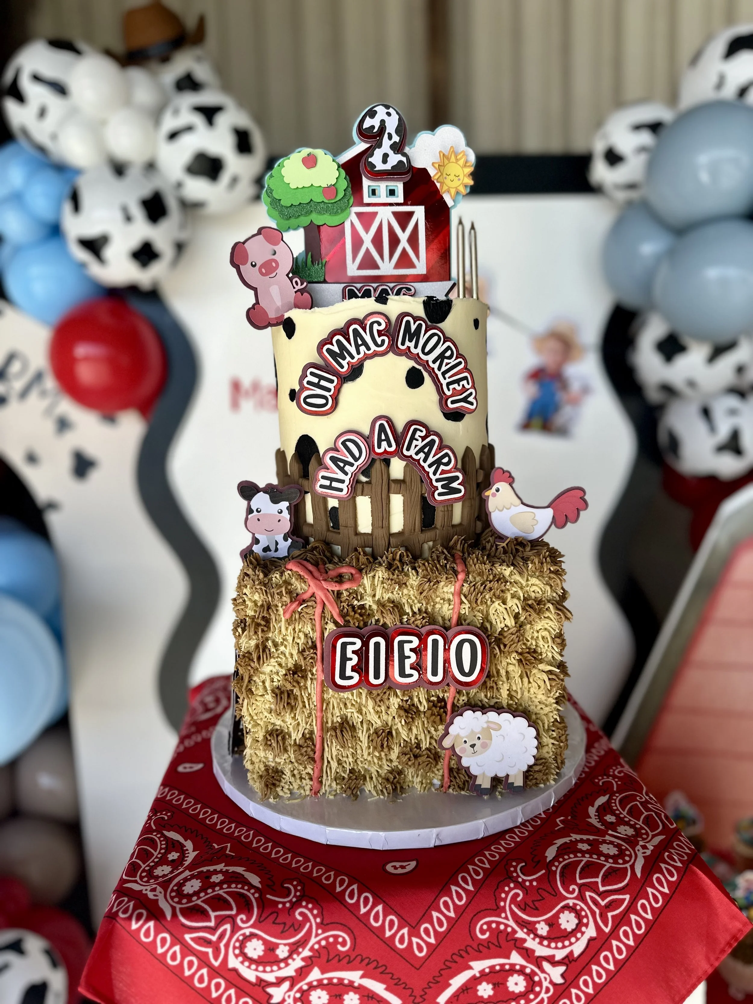 A farm-themed birthday cake decorated with farm animal toppers, including a pig, a chicken, and a sheep. The cake has multiple tiers with farm-related decorations. The cake is placed on a red bandana-patterned cloth.