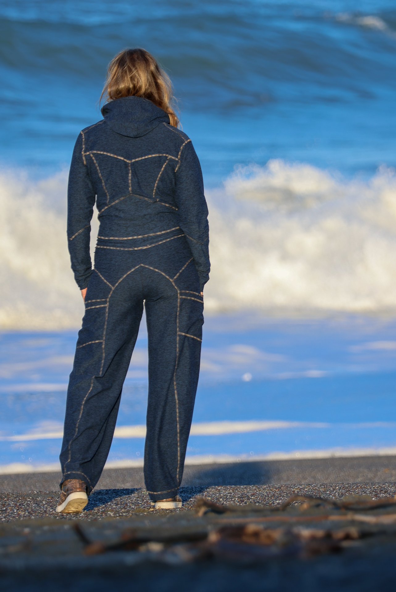 Person with long hair wearing a dark blue hoodie and loose jeans, standing on a beach facing the ocean waves.