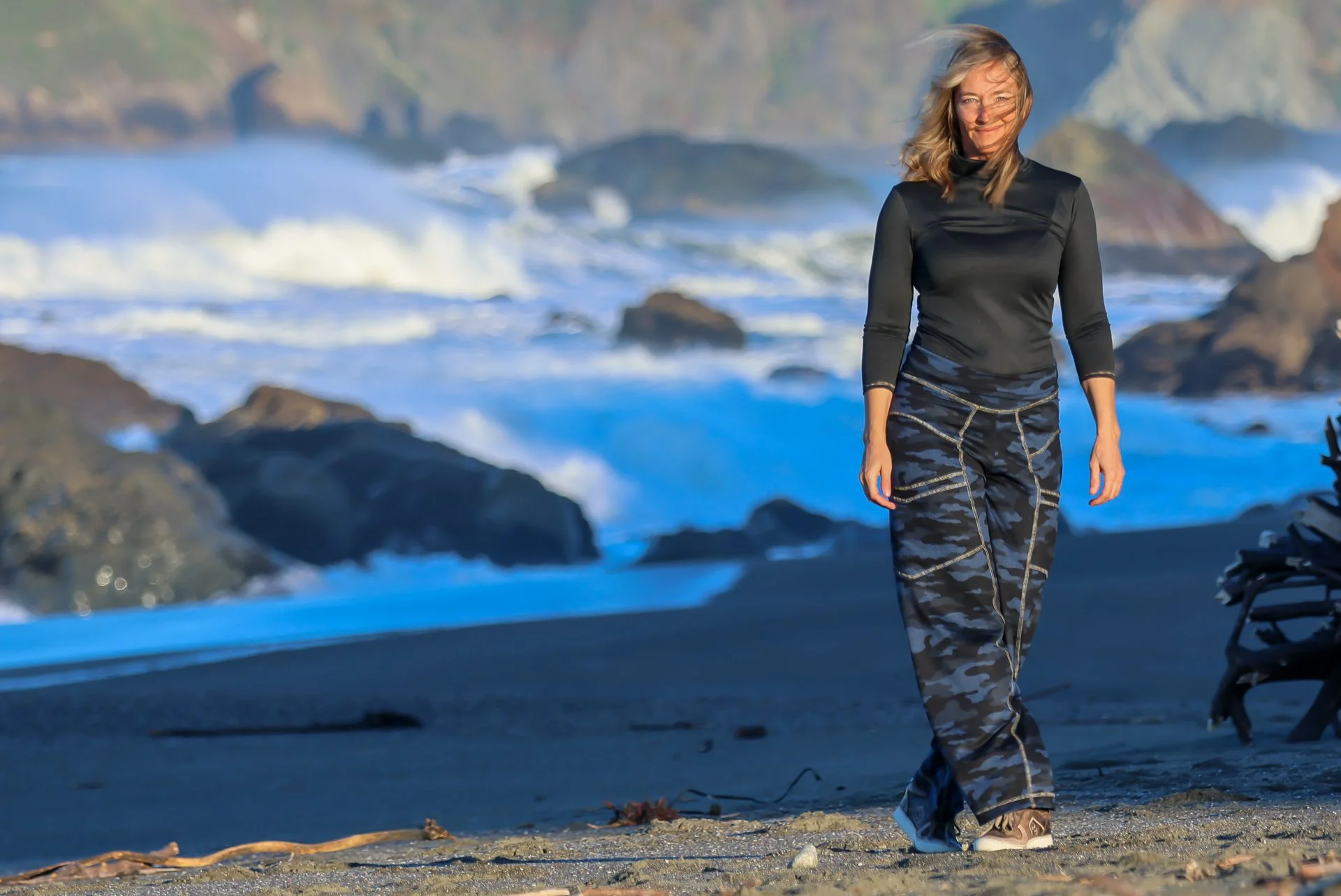 A woman walking on a beach with rocky shoreline and ocean waves in the background.