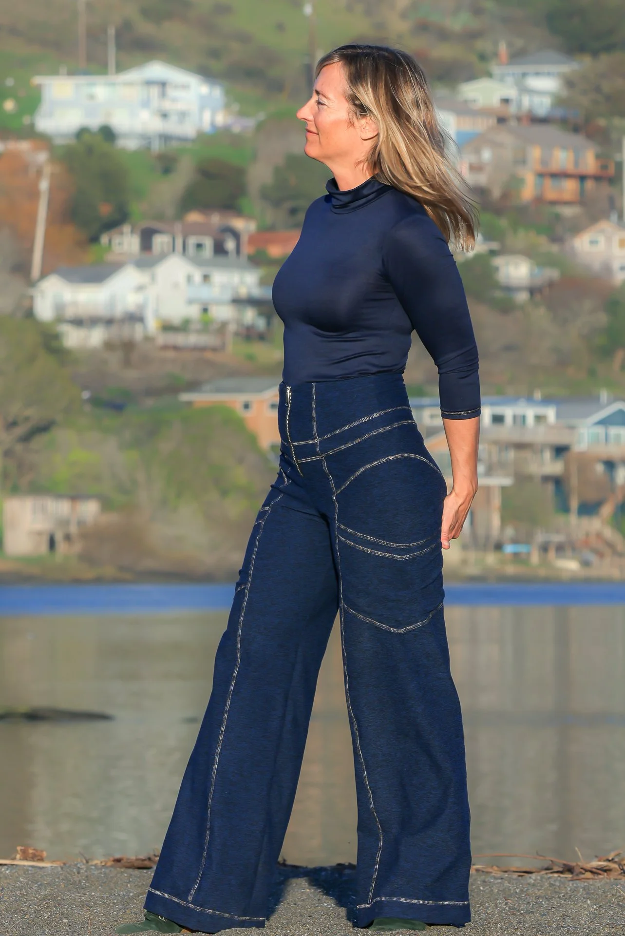 Woman in a navy blue turtleneck top and wide-leg jeans standing outdoors near water with homes on a hillside in the background.