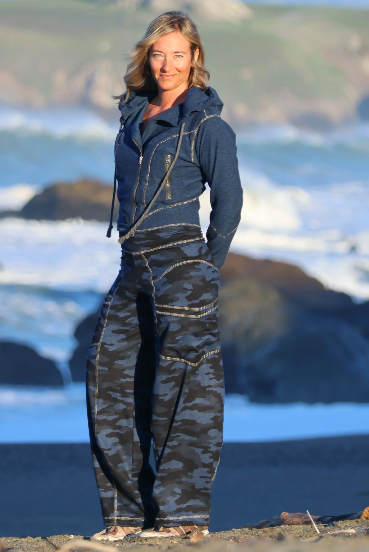 Woman standing on a beach near rocks with ocean waves in the background, wearing a Midnight Blue Bomber Jacket and Midnight Blue Camo Grand Daphne Pant.
