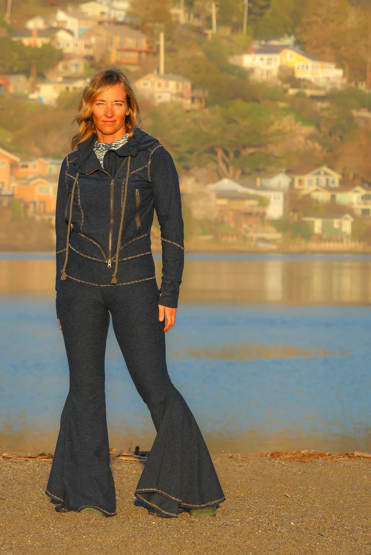 A woman, wearing Midnight Blue Hope Pants and a Midnight Blue Bomber Jacket, standing on the beach near a lake with colorful houses on a hillside in the background during sunset.