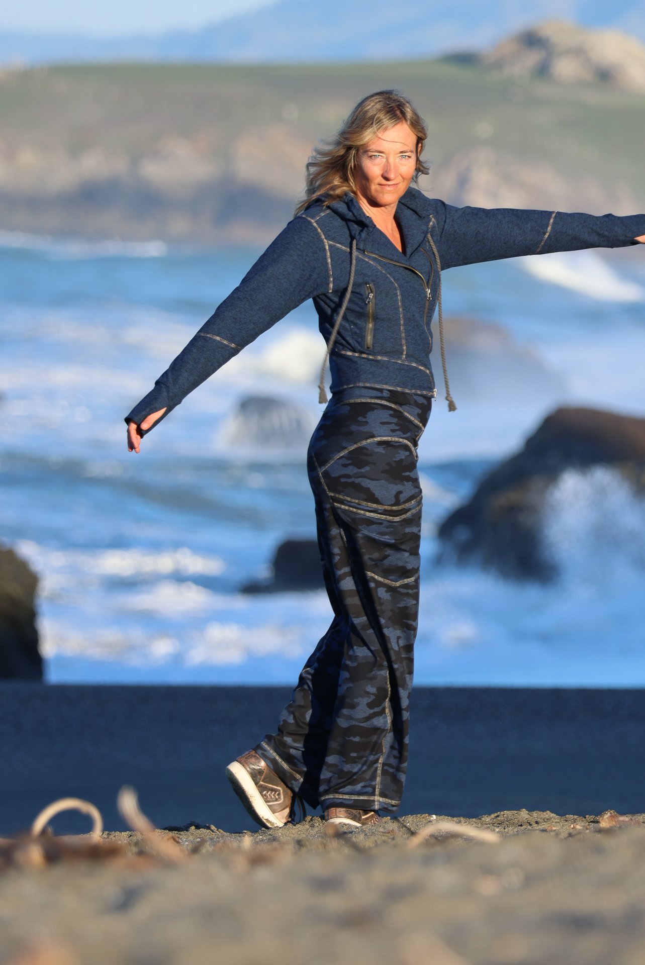 Woman standing on sandy beach with ocean and rocks in background, arms outstretched, wearing a hoodie, camouflage pants, and sneakers.