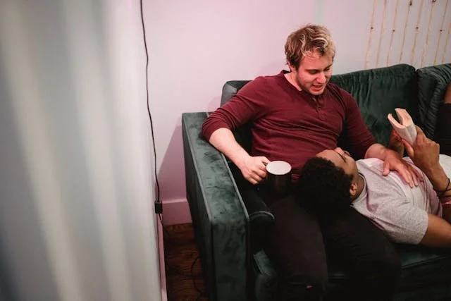 Interracial LGBT couple relaxing at home, reading and enjoying coffee together on a green sofa.