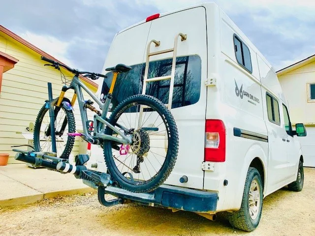 White van with rear bike rack carrying two bicycles, parked near a house.