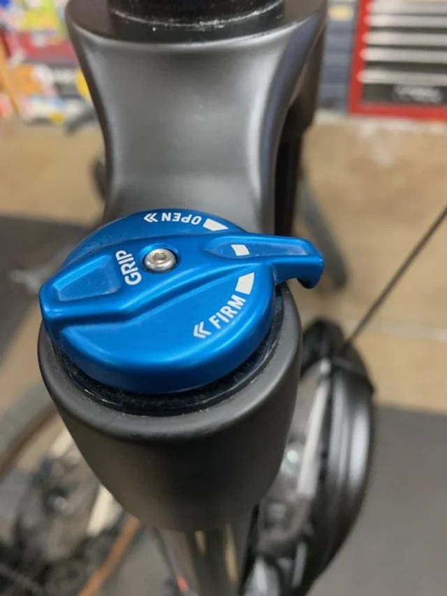 Close-up of a blue bike suspension fork with grip adjustment knob labeled "FIRM" and "OPEN."