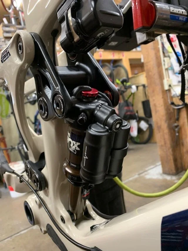 Close-up of a bicycle's rear suspension system, featuring a Fox shock absorber, set in a workshop with tools and other bikes visible in the background.