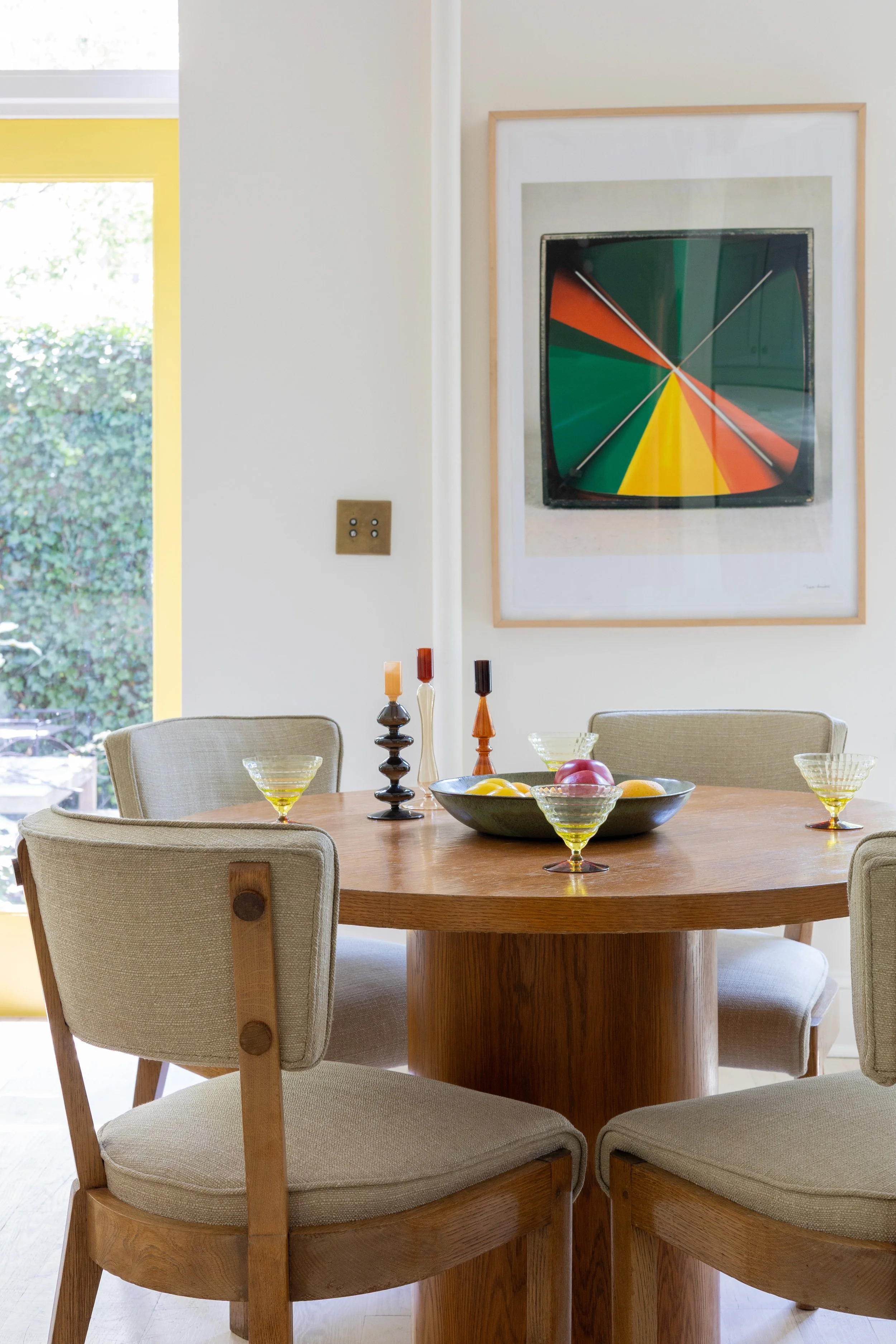 Dining room with a round wooden table set with glasses, a bowl of fruit, and decorative candlesticks, with a large colorful abstract painting on the wall and a window in the background.