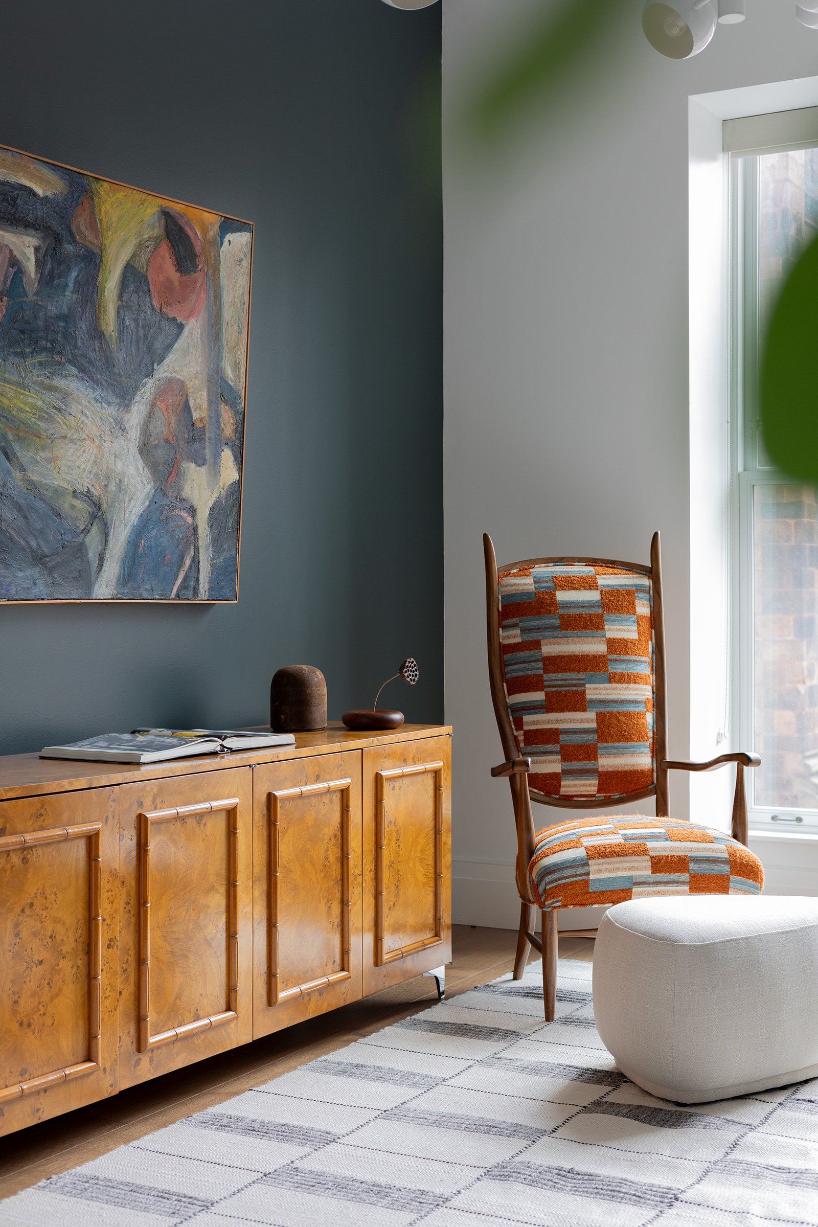 Interior of a modern living room featuring a wooden sideboard with decorative objects, a colorful patterned armchair, a white ottoman, a large abstract painting on the wall, and a window letting in natural light.