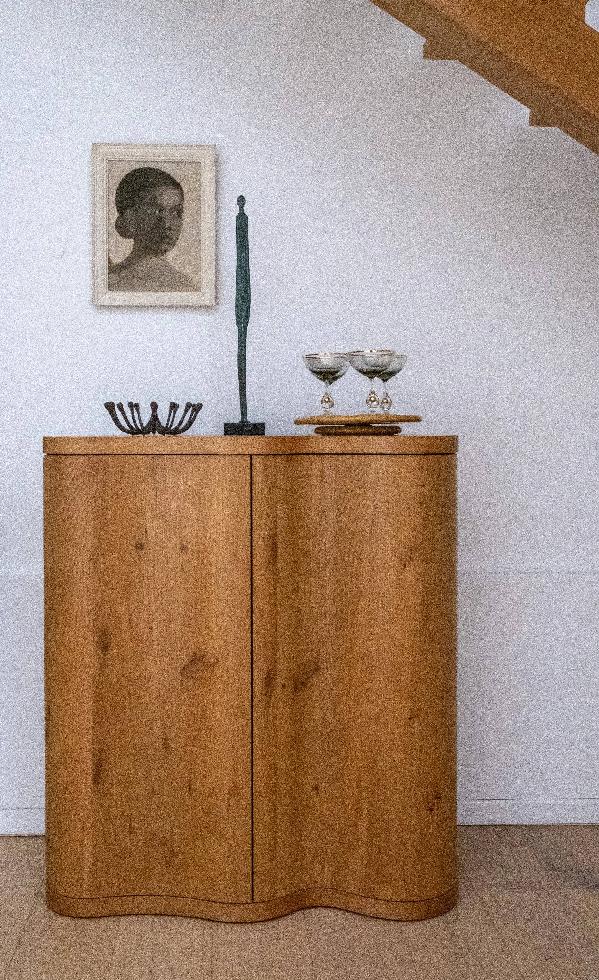 A wooden cabinet on a light-colored wooden floor. On top of the cabinet are three empty cocktail glasses on a wooden tray, a decorative black metal object, and a tall, thin, greenish sculpture. Above the cabinet hangs a framed black-and-white photograph of a woman with short hair, looking to the side.
