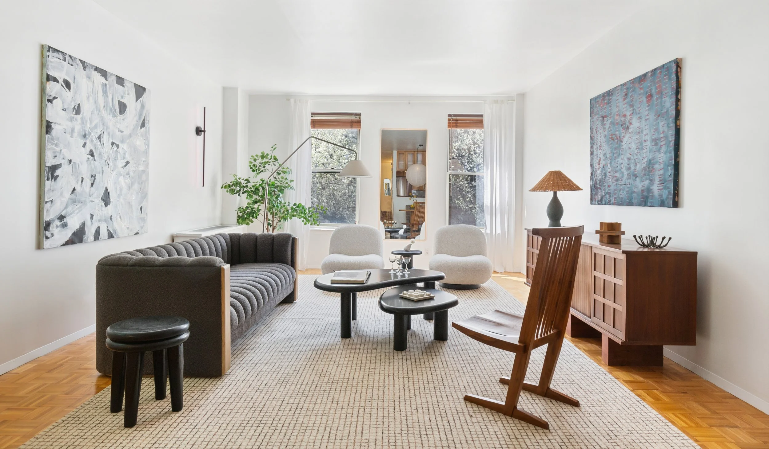 Living room with two white armchairs, a gray tufted sofa, black oval coffee tables, and a wooden rocking chair. Large window with white curtains, wooden floor, and abstract paintings on white walls.