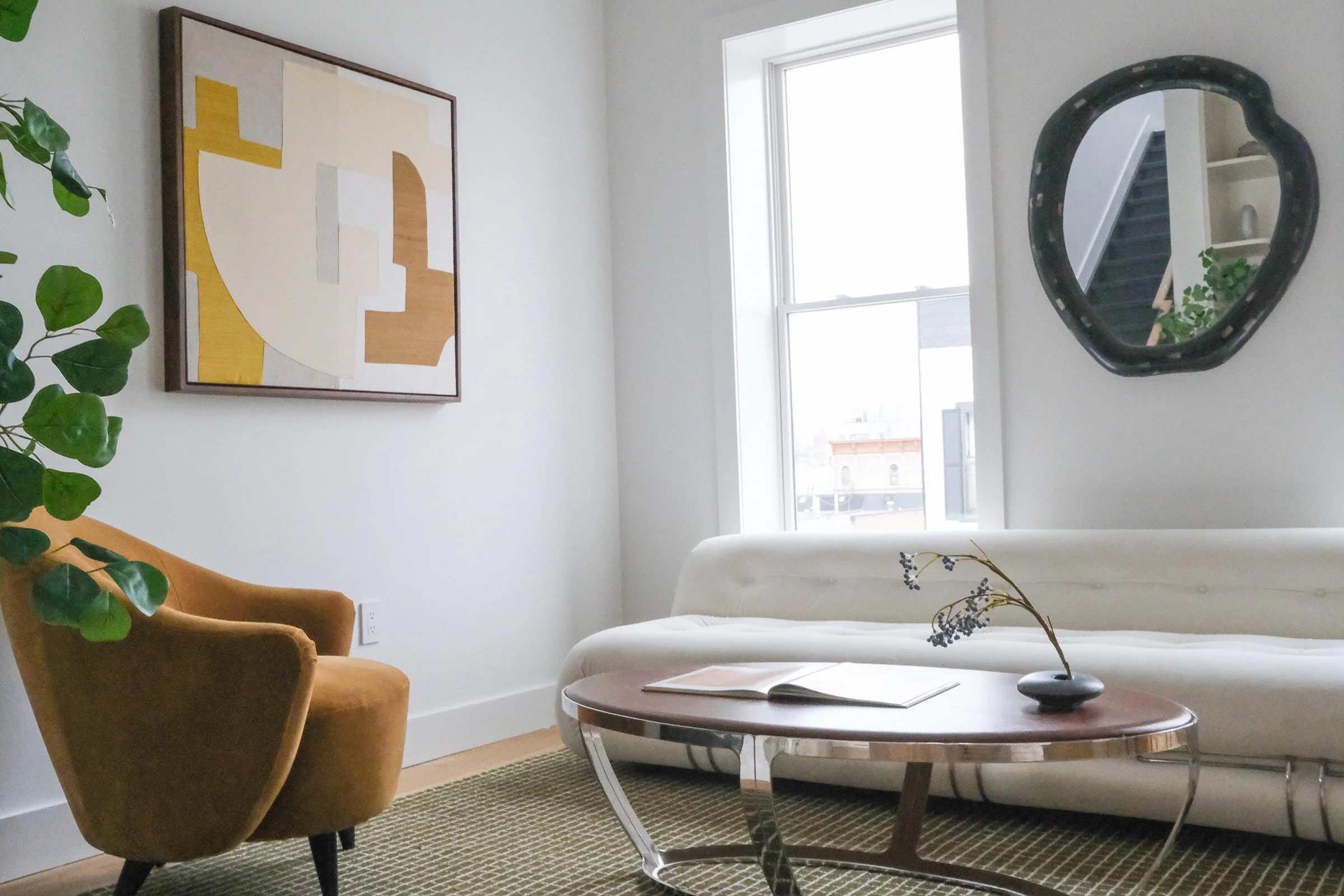 A living room with a mustard-colored armchair, a white sofa, a round wooden coffee table with a vase and open book, a large window letting in natural light, an abstract painting, a black framed mirror, and a green plant.