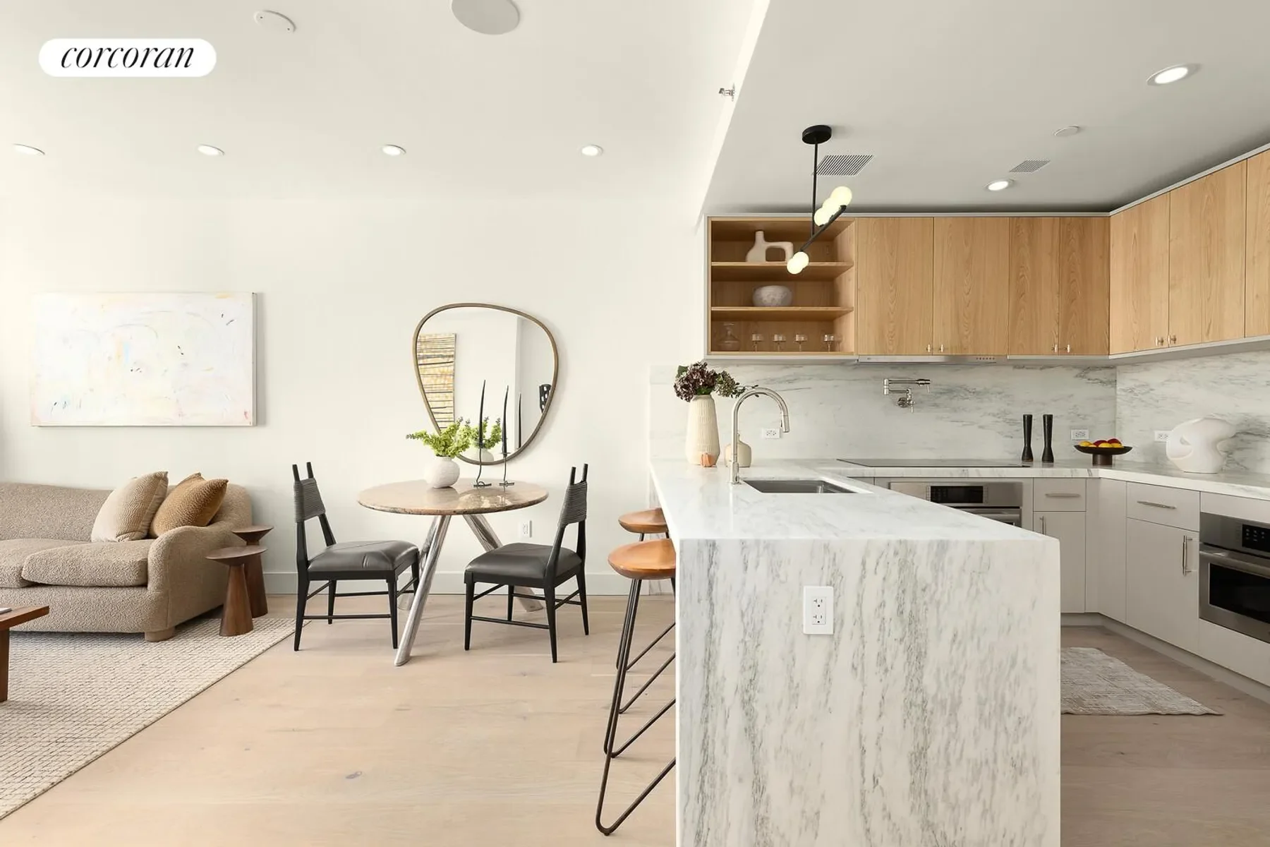 Open-concept living space with a beige sofa, a round dining table with two black chairs, and a kitchen with white cabinets, a marble countertop, and wooden upper cabinets. Decorative items include a mirror, vases, and a bowl with fruit.