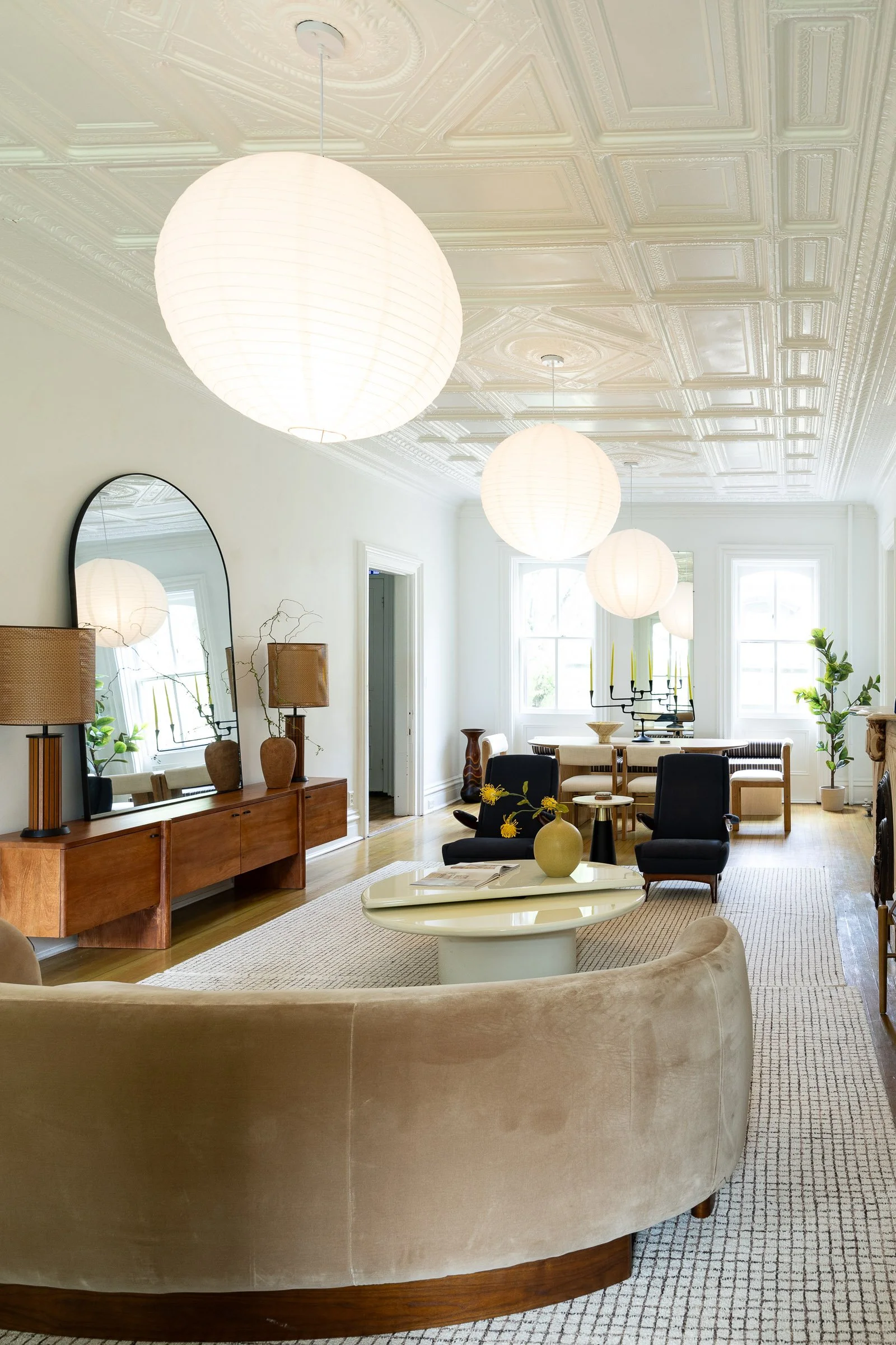 Brightly lit living room with white walls, ornate ceiling, and large paper lantern pendant lights. Features a curved beige sofa, black armchairs, a white coffee table with a vase, a wooden sideboard with lamps and a mirror, and a dining area with a white table, built-in bench, and candle holders, decorated with plants.