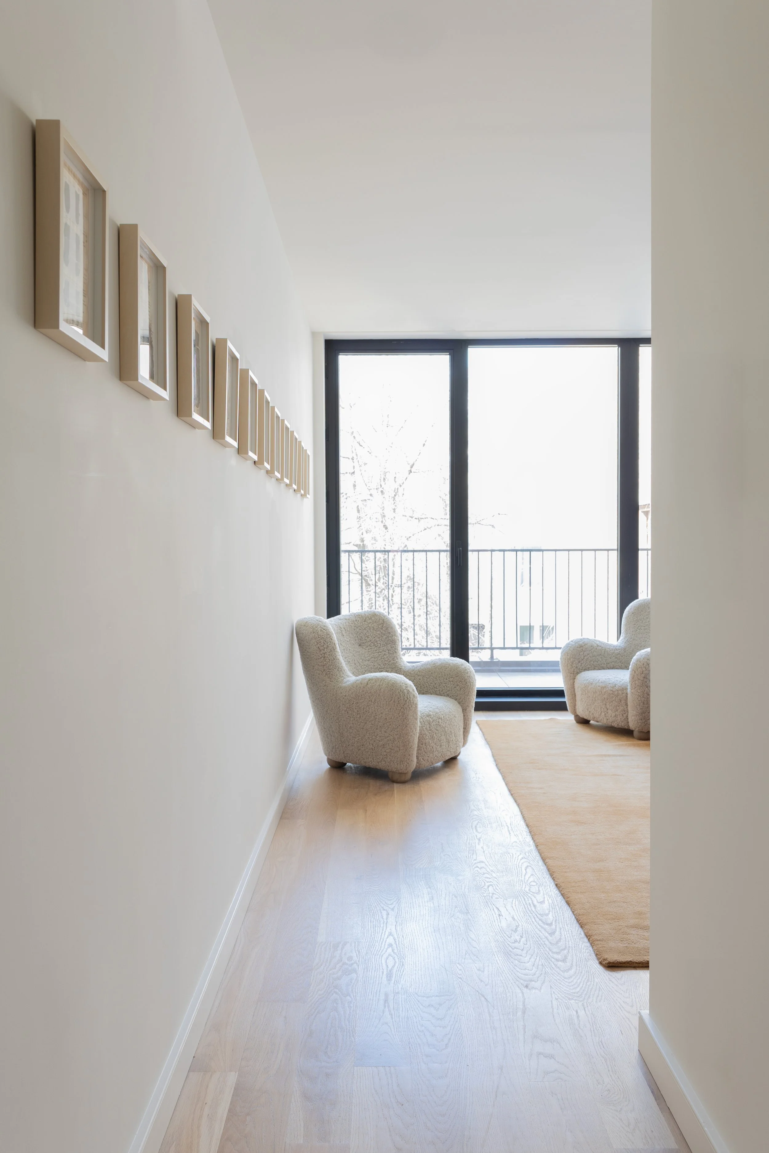 Living room with two cream-colored armchairs, framed artwork on the wall, wooden floor, and glass sliding doors leading to a balcony with snow outside.