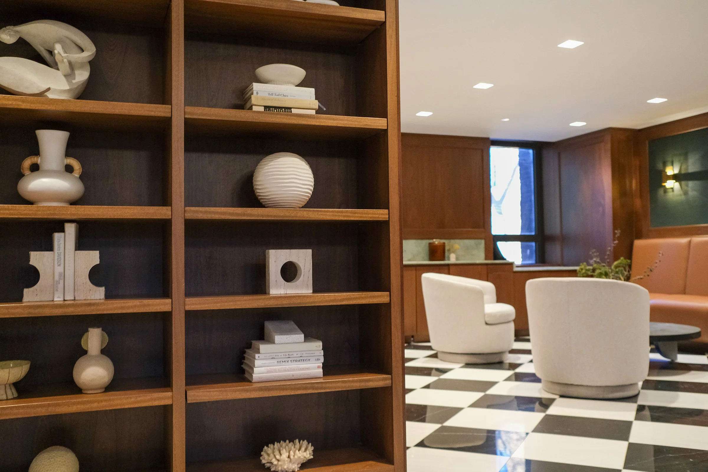 Interior of a modern living room with a wooden bookshelf filled with neutral-colored decorative objects and books, a checkered black and white tiled floor, and seating arrangements including white and beige chairs and a peach sofa, with a window and green wall with a sconce light in the background.