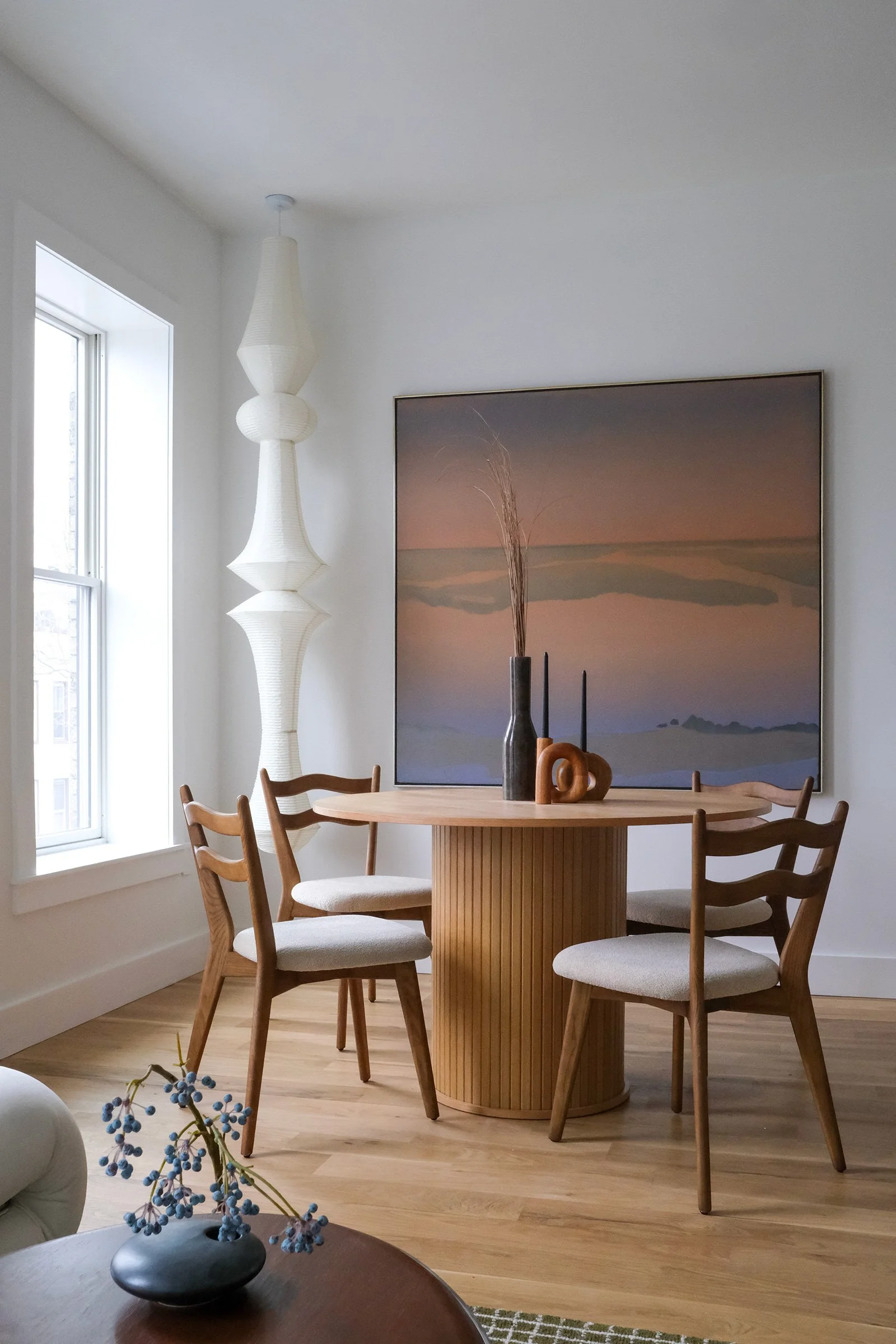 A dining room with a round wooden table and four chairs, a large abstract painting on the wall, a window letting in natural light, and decorative vases and candles on the table.